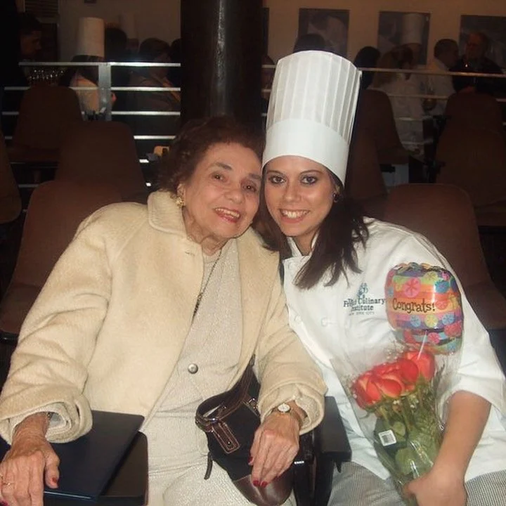 An elderly woman and a young woman in a chef's uniform, including a tall chef's hat, sitting together in a restaurant. The young woman is holding a bouquet of red roses and a 'Congrats' balloon.