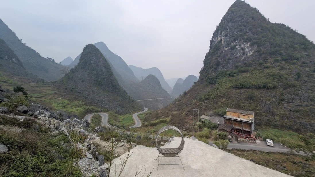 Winding mountain road through lush green hills with a building and parked car at the base, and foggy mountain peaks in the background.