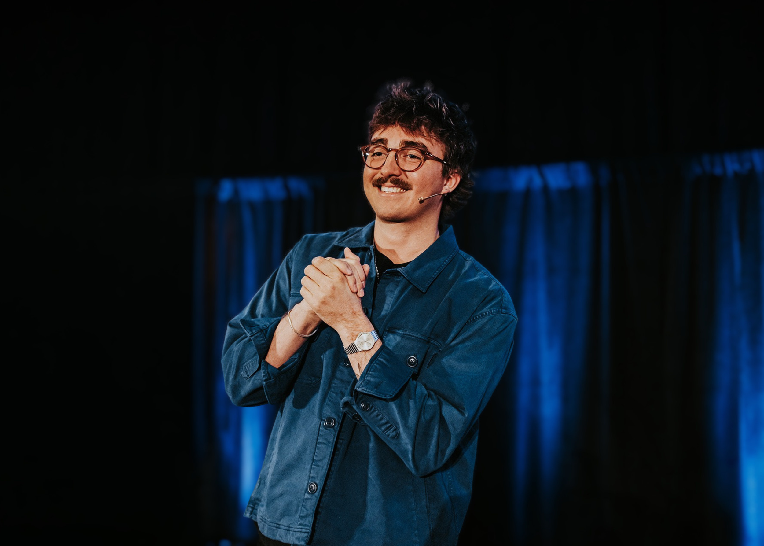 A man with glasses and a mustache on stage, smiling with hands clasped, wearing a denim jacket and a watch, with dark curtains and blue lighting in the background.
