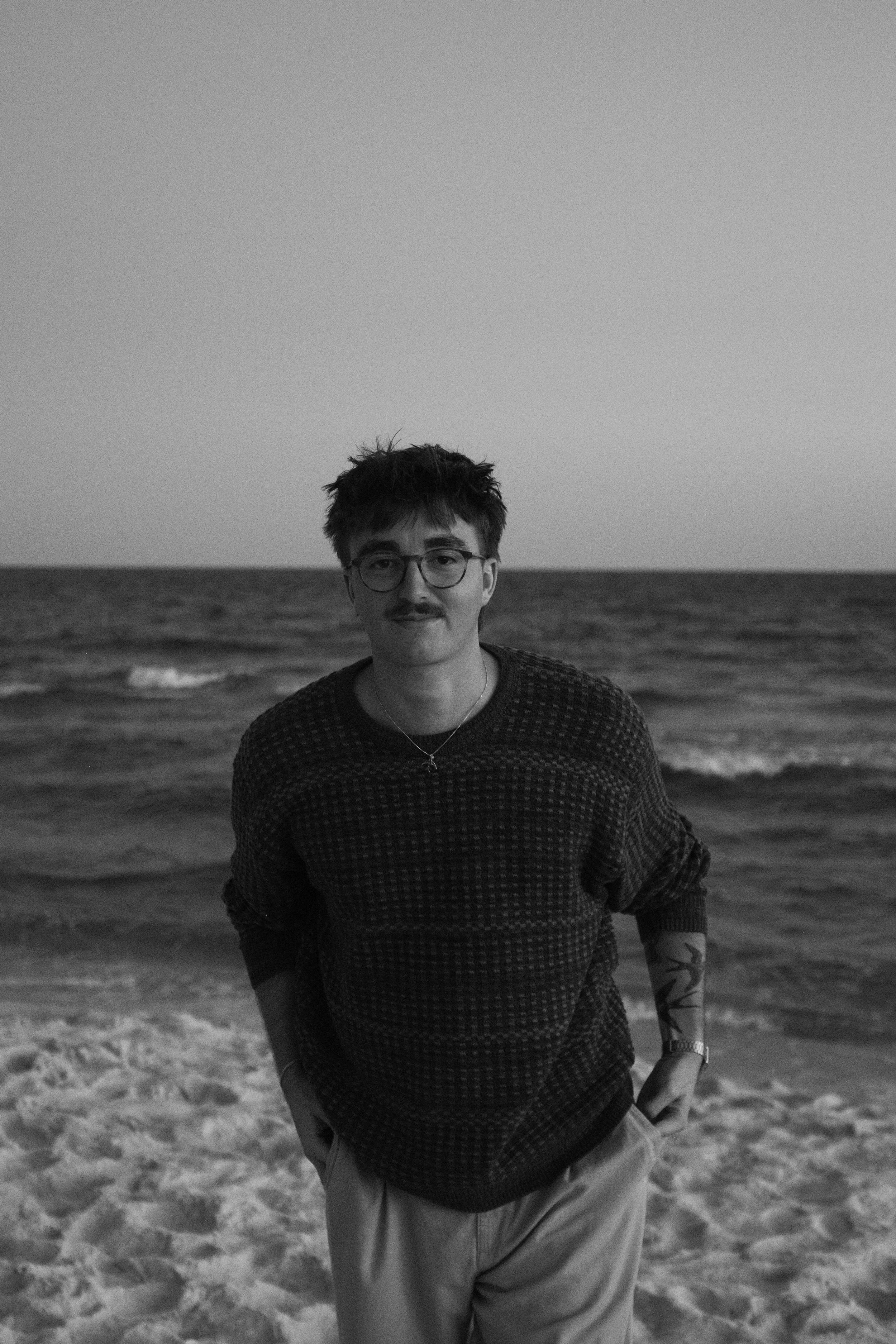 A young man with glasses and a mustache standing on the beach with the ocean in the background.