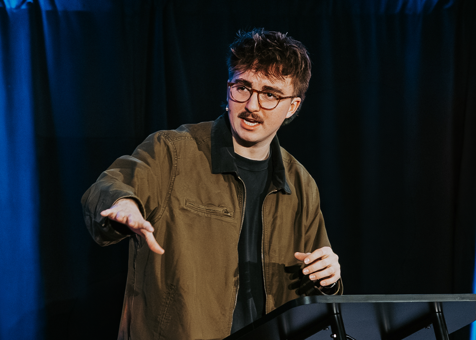 A man with glasses and a mustache, wearing a brown jacket and black shirt, speaking or gesturing during a presentation or performance on stage with dark curtains in the background.