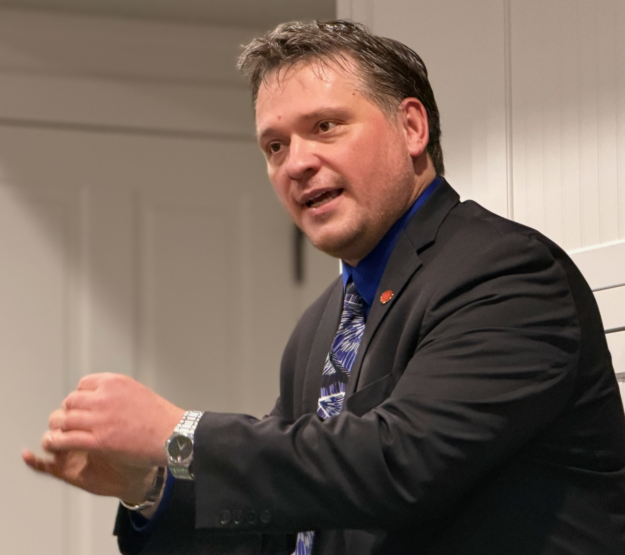 A man in a suit and blue shirt speaking and gesturing in front of a plain background while talking about coin magic..