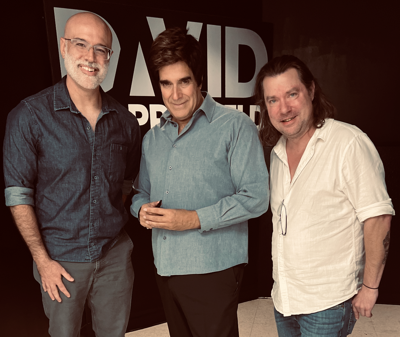 Three men standing together in front of a black wall with large white text. The man on the left has a beard, glasses, and is wearing a dark denim shirt. The man in the middle has dark hair, is wearing a light blue shirt, and is holding a pen. The man on the right has long hair, is wearing a white shirt, and glasses hanging from his collar.