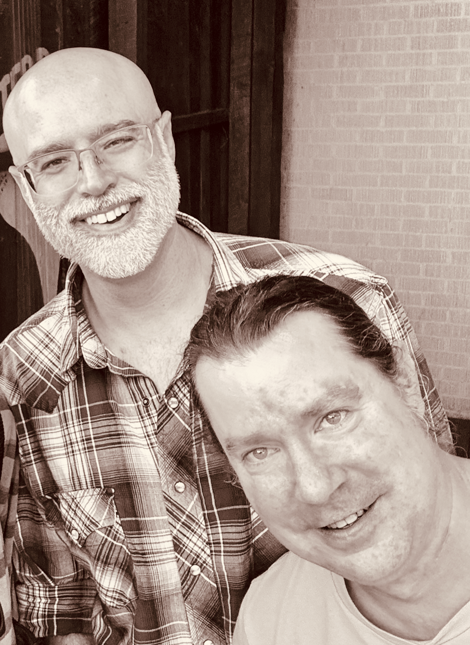 Two smiling men, one with a beard and glasses wearing a plaid shirt, Joshua Glantzberg, and the other with long hair pulled back, and no glasses wearing a light-colored shirt, J Weldon, taking a selfie together outside.