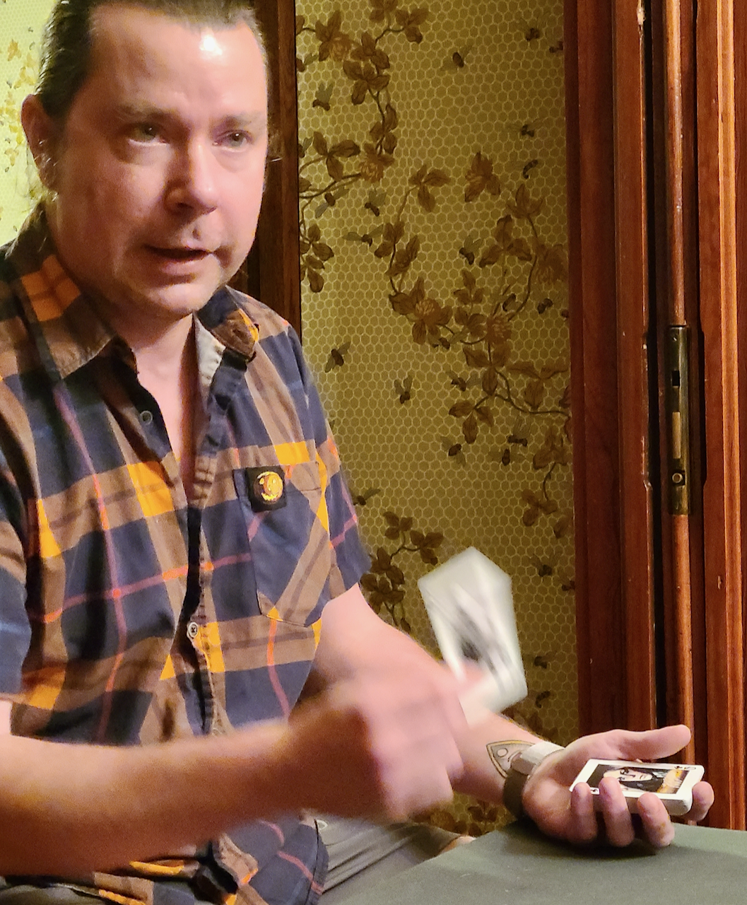 A man with short, dark hair, wearing a plaid shirt, appears to be in the middle of dealing or shuffling money and holding a phone, with a background that has a patterned wallpaper with leaves.