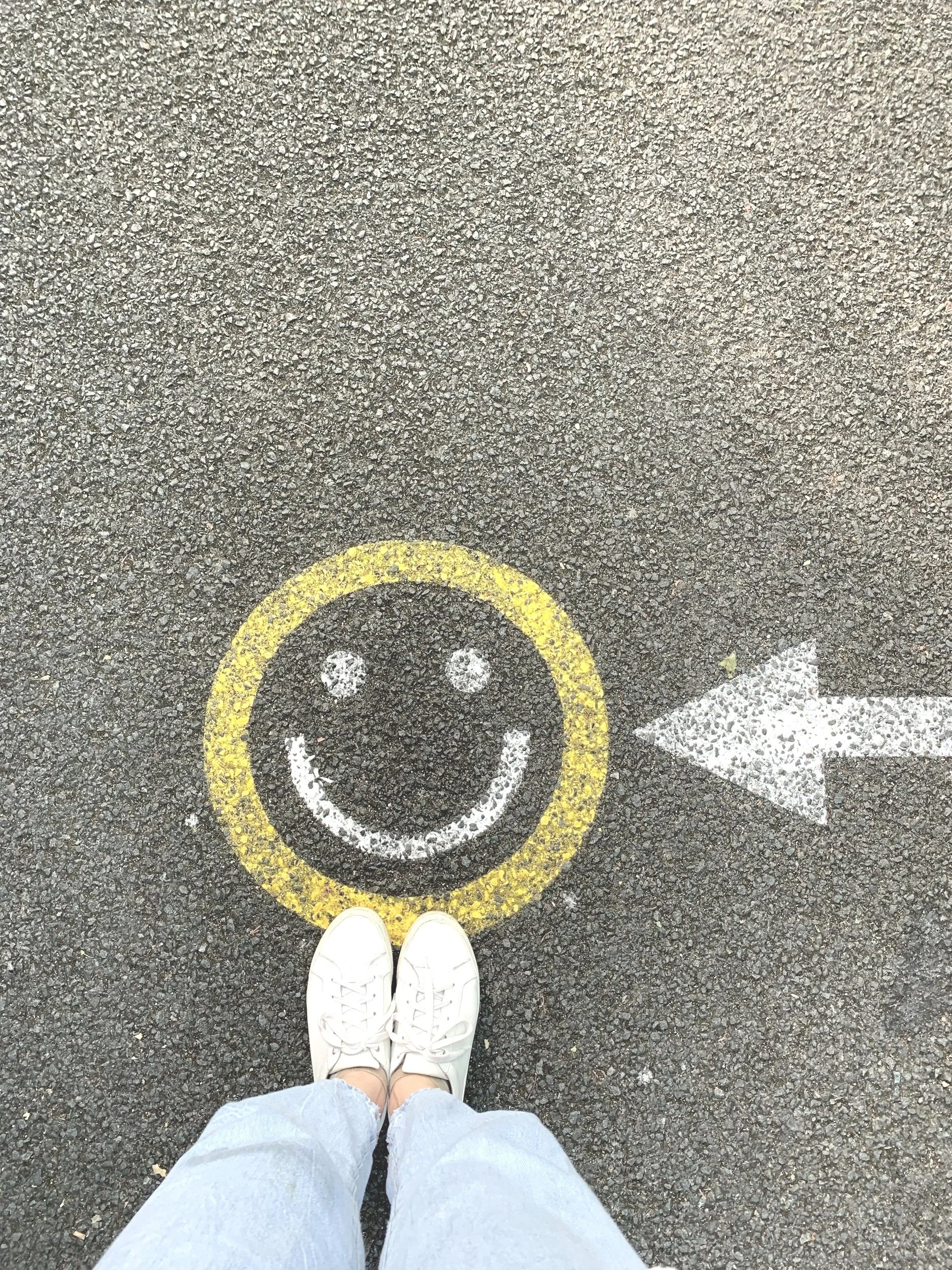 Photo of a person's feet wearing white sneakers and light jeans standing on pavement marked with a yellow smiley face and a white arrow pointing to the right.
