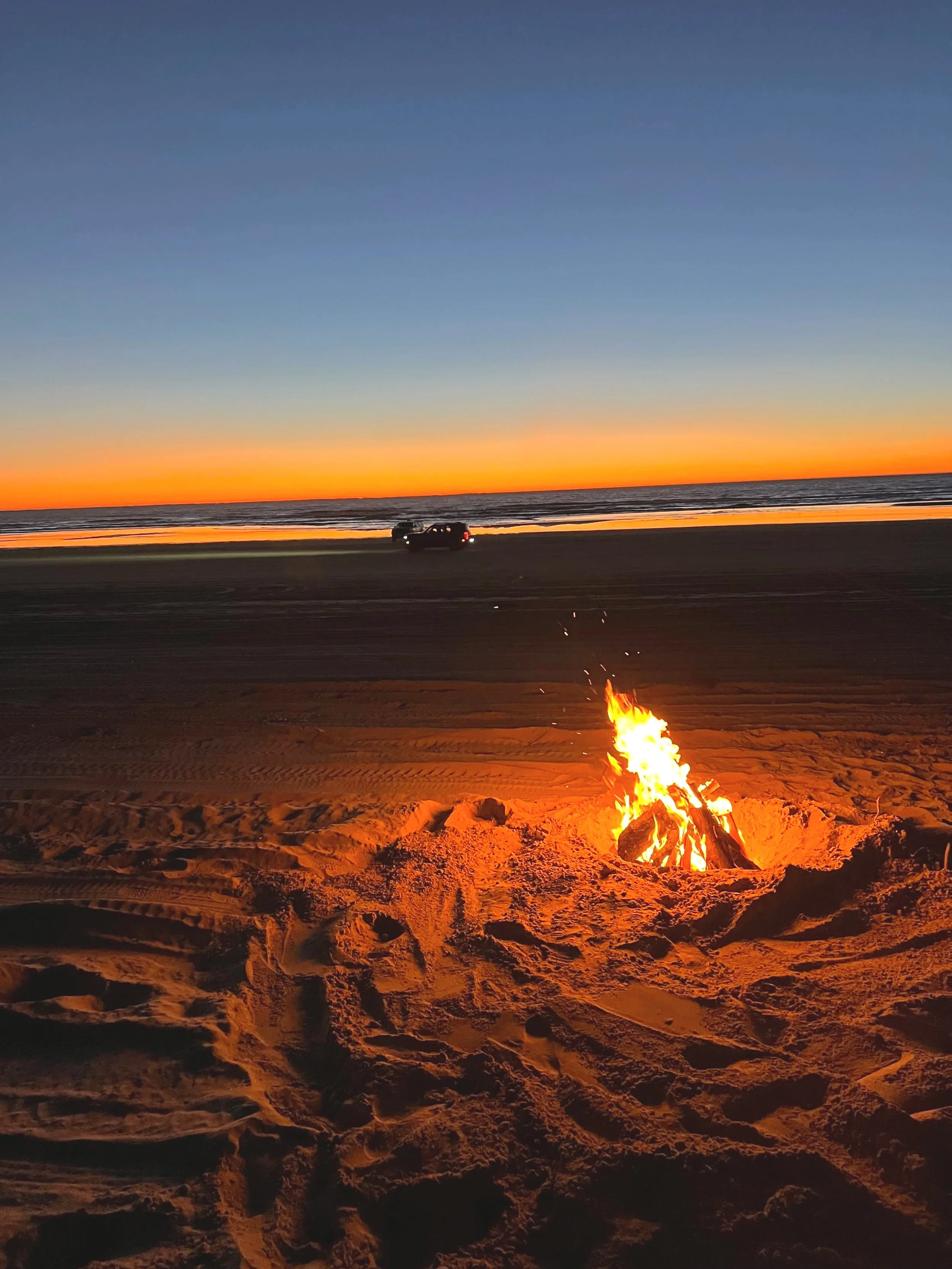 Beach at sunset with a bonfire in the foreground and a vehicle near the water in the background.