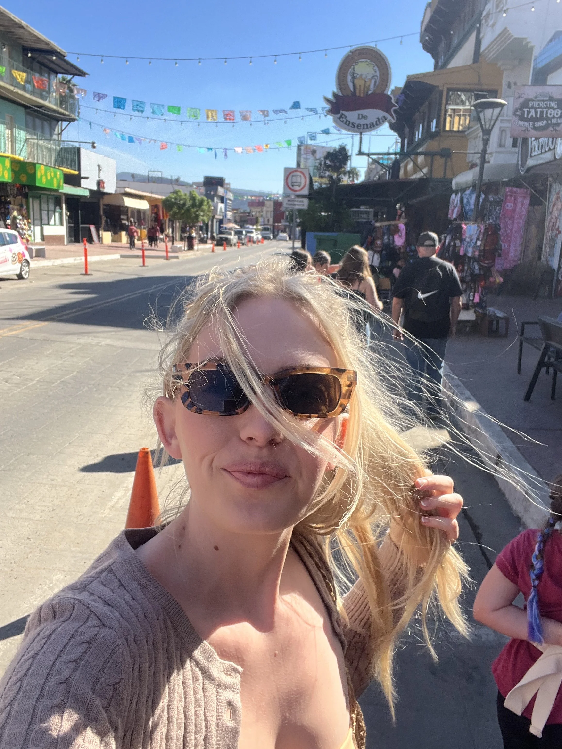 Woman with blonde hair and sunglasses taking a selfie on a busy street with shops and colorful banners overhead.