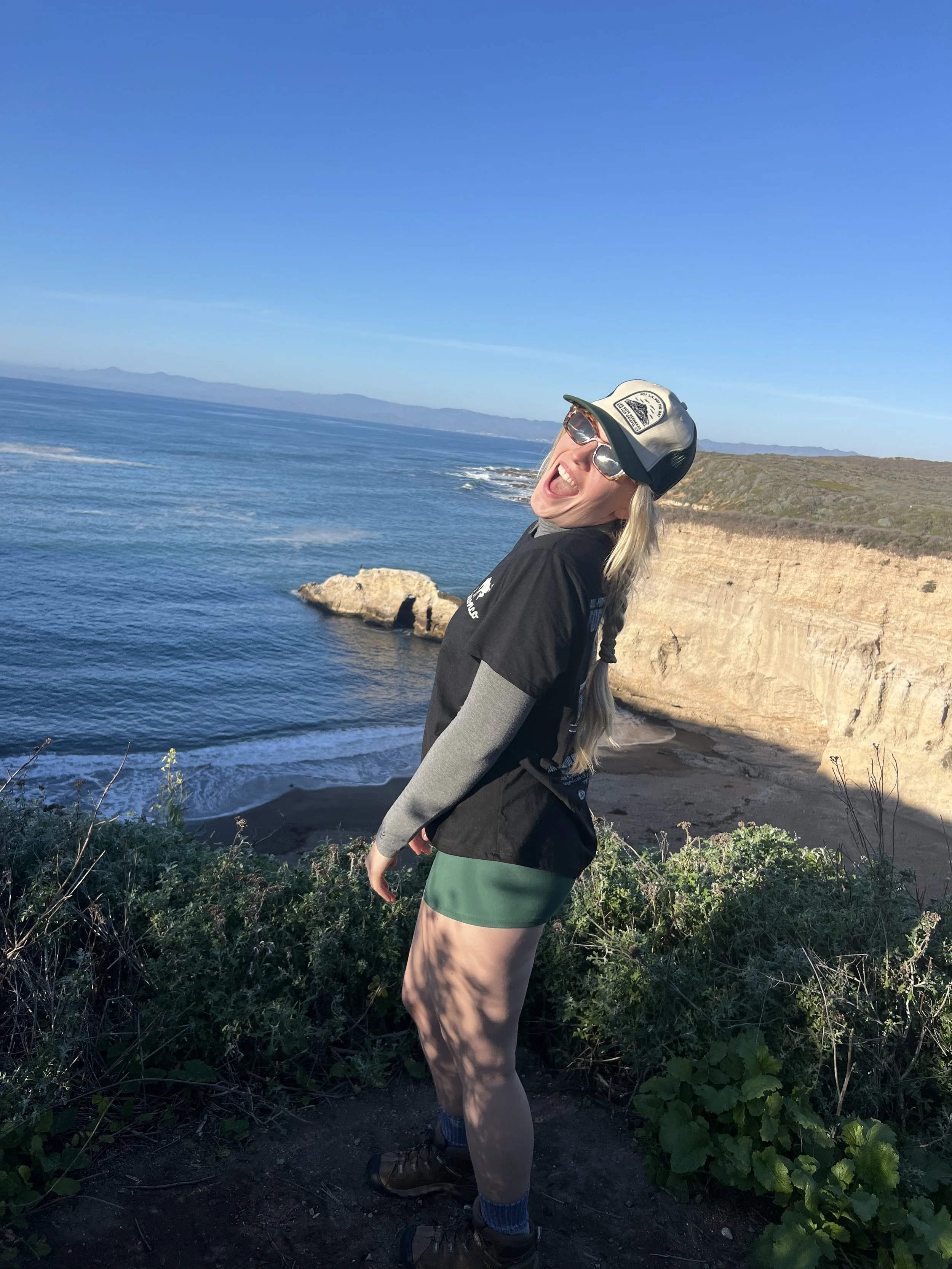 A woman with long blonde hair, wearing sunglasses, a cap, a black and gray jacket, green shorts, and hiking boots, standing on a cliff overlooking the ocean with rocky shoreline and a clear blue sky.