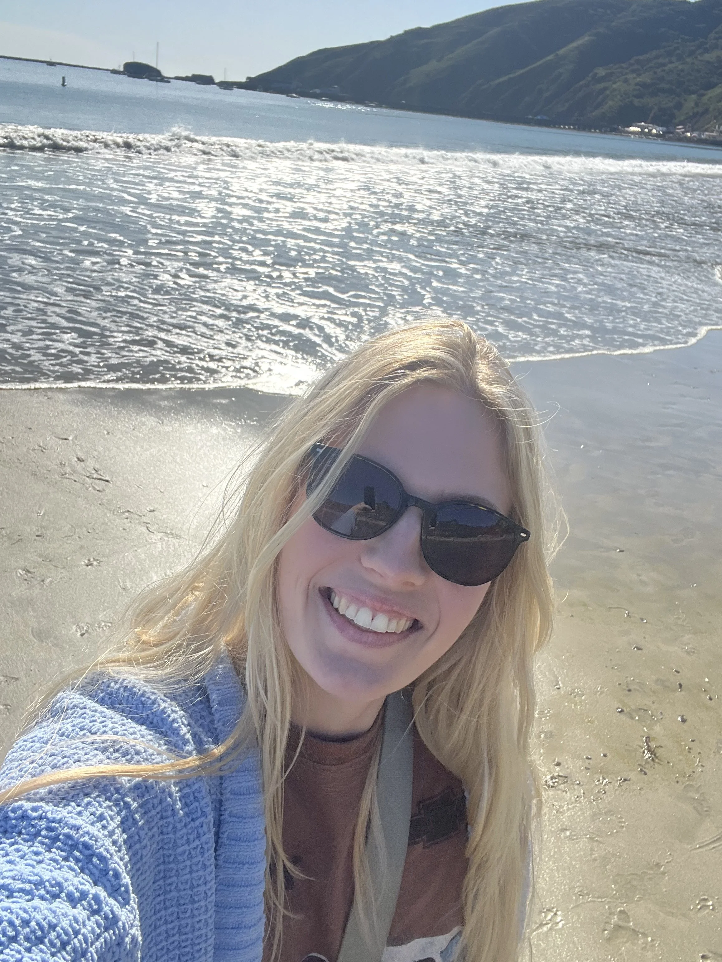 A woman with blonde hair wearing sunglasses taking a selfie on a beach, with the ocean and green hills in the background on a sunny day.
