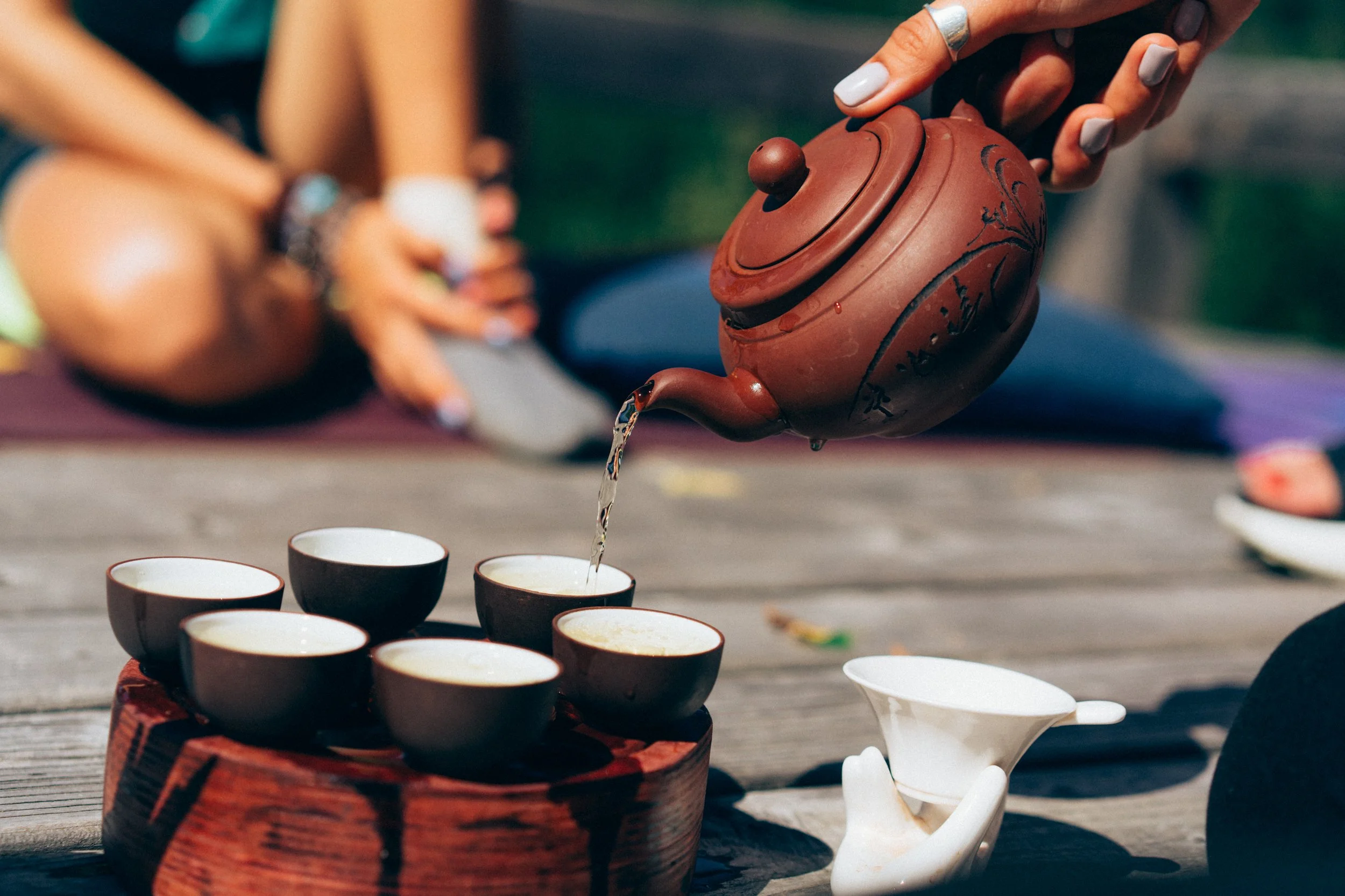 Person pouring tea into small cups on a wooden table outdoors with people sitting on a bench in the background.