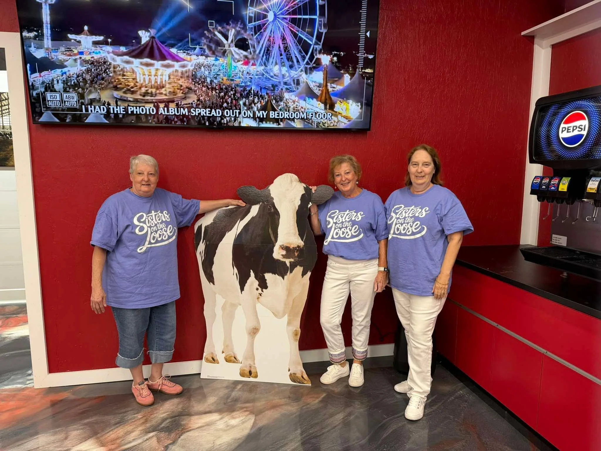 Three women standing next to a cardboard cutout of a cow at an amusement park. Behind them, a large screen shows a night scene of a carnival with a Ferris wheel and other rides. The women are smiling, and the women and the cutout are dressed in blue shirts that say 'Sisters on the Loose'.