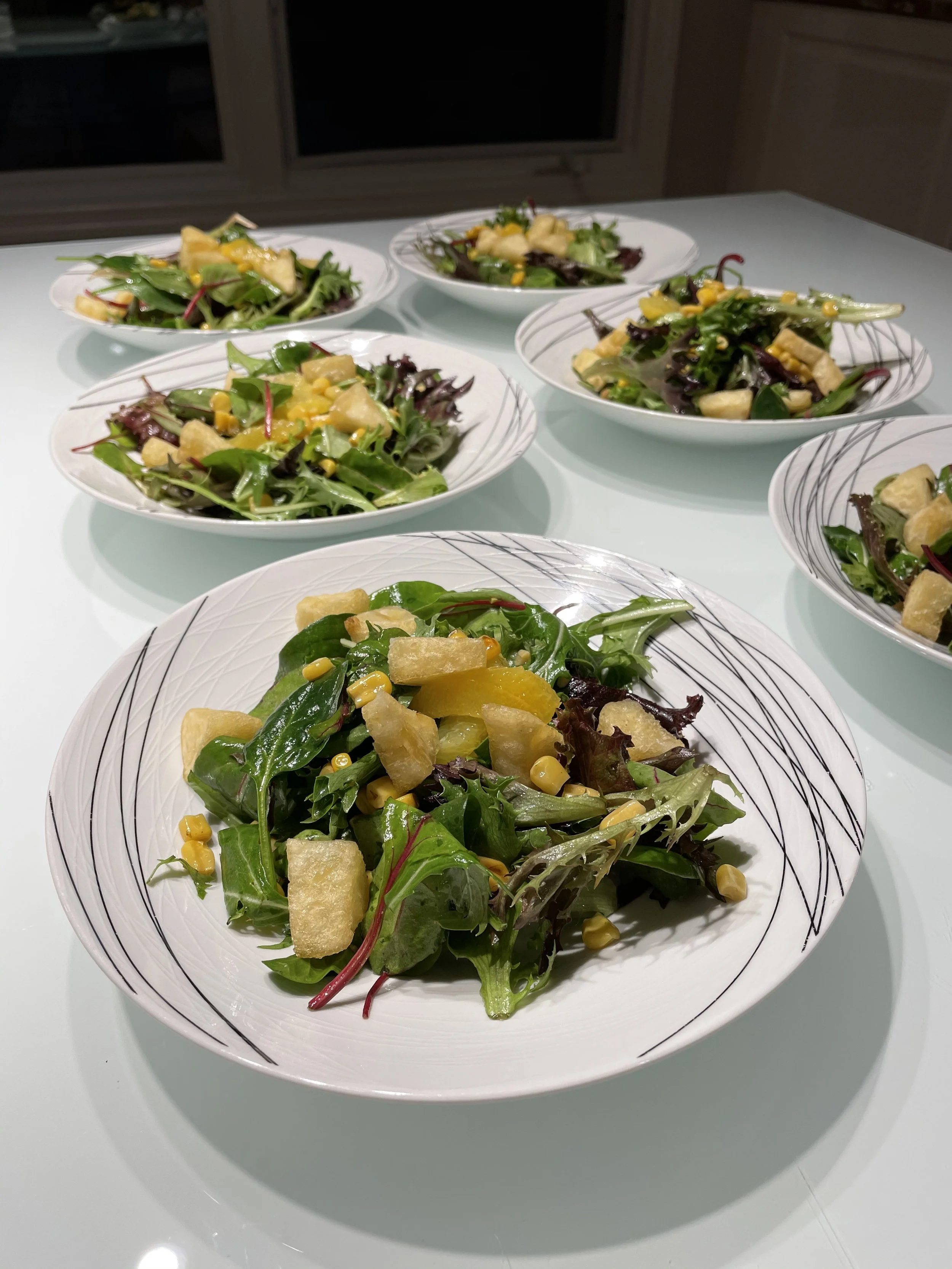 Plates of mixed greens salad with corn, cheese, and croutons on a white table, with multiple servings.
