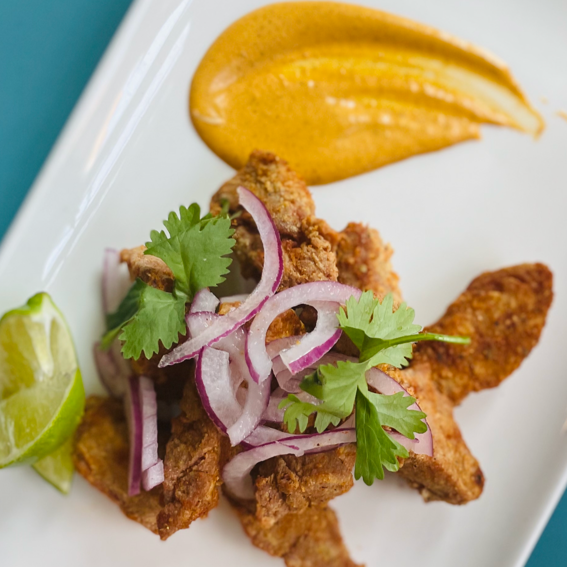 A plate of fried fish garnished with cilantro, red onion slices, and lime wedges, served with a dollop of mustard sauce.