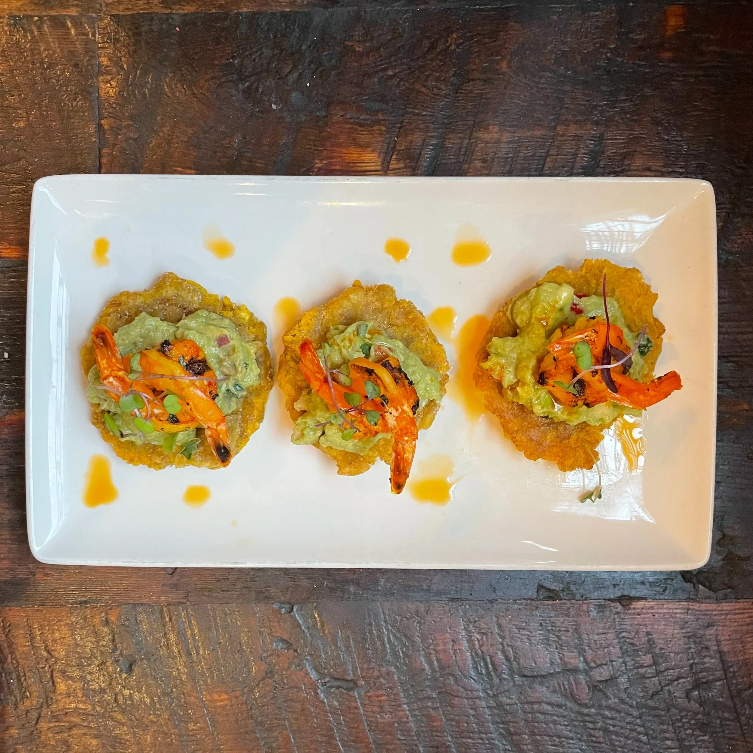 Three pieces of fried food topped with guacamole, shrimp, and microgreens on a white rectangular plate with a drizzle of sauce, set on a wooden table.