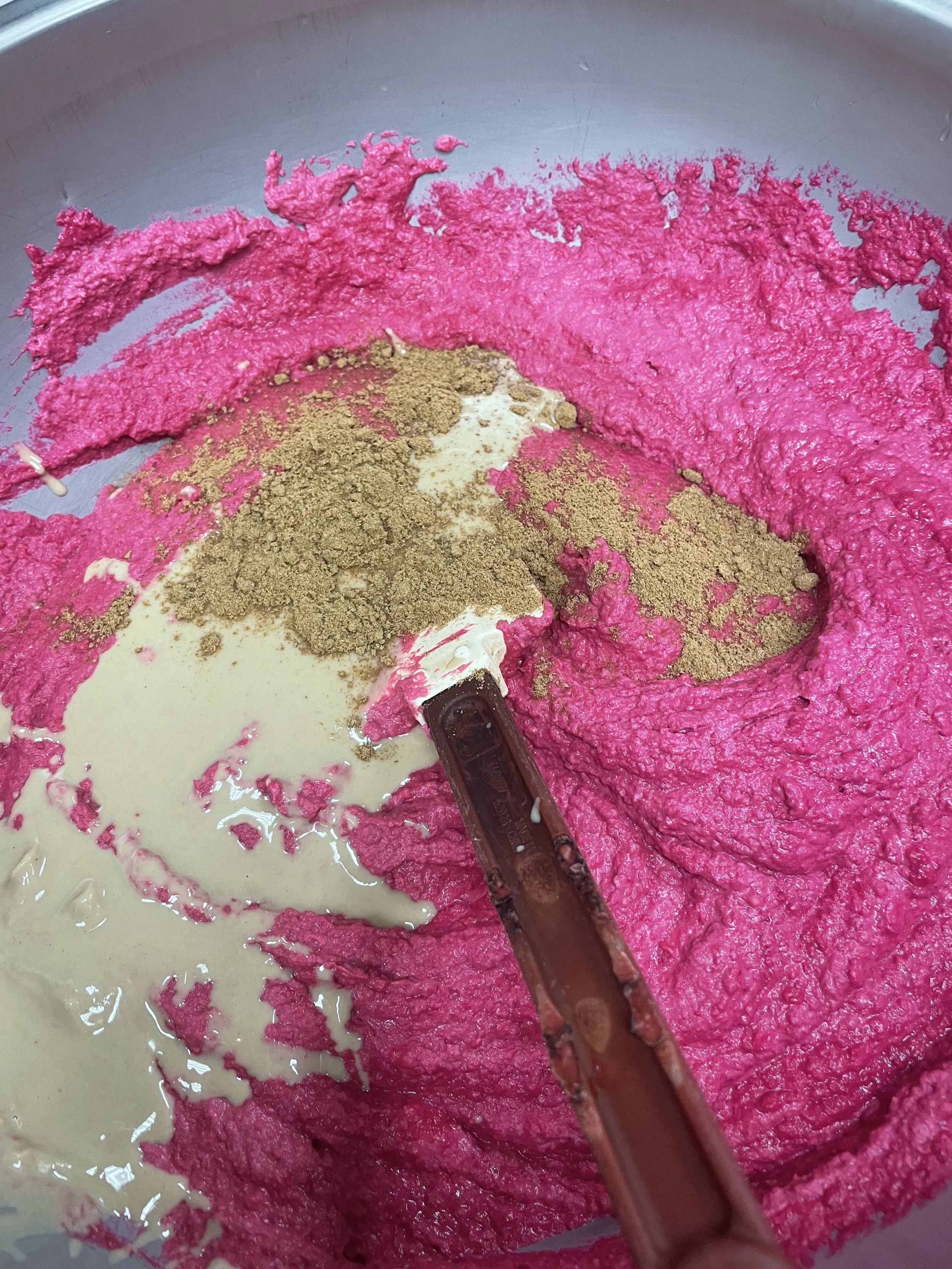 A mixing bowl containing pink batter, beige powder, and liquid, with a spatula mixing the ingredients.
