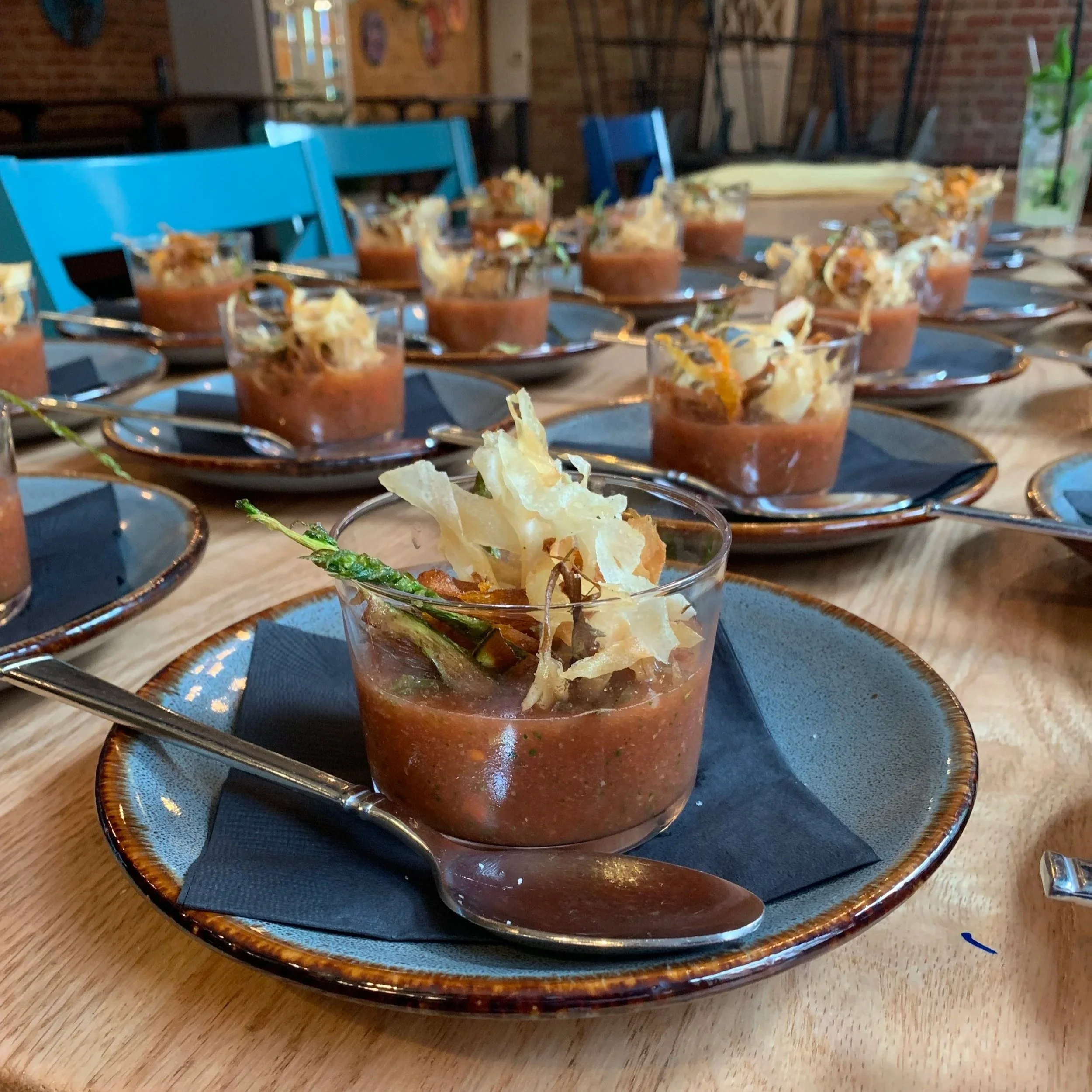 Multiple small glasses filled with a red-brown soup topped with crispy cheese chips and a green garnish, arranged on a wooden table with dark plates and silver spoons.
