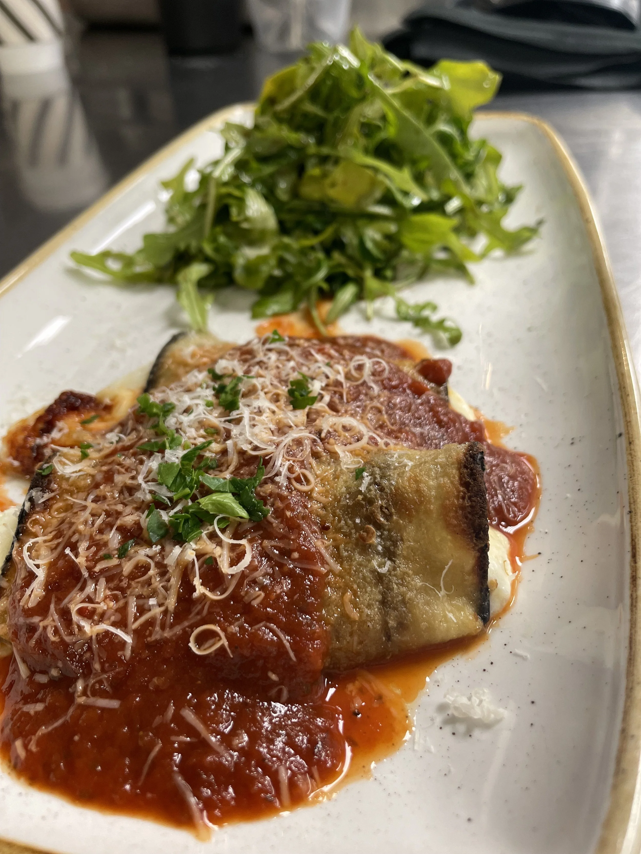 Baked eggplant parmesan with melted cheese, tomato sauce, and chopped basil, served with a side of fresh greens on a white plate.