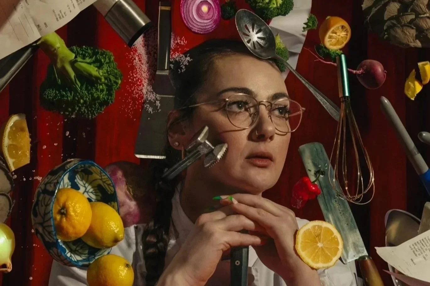 Chef Jessica Baston with glasses rests her chin on her hands at a table surrounded by various kitchen utensils and food items, including lemons, broccoli, a whisk, and a grater.