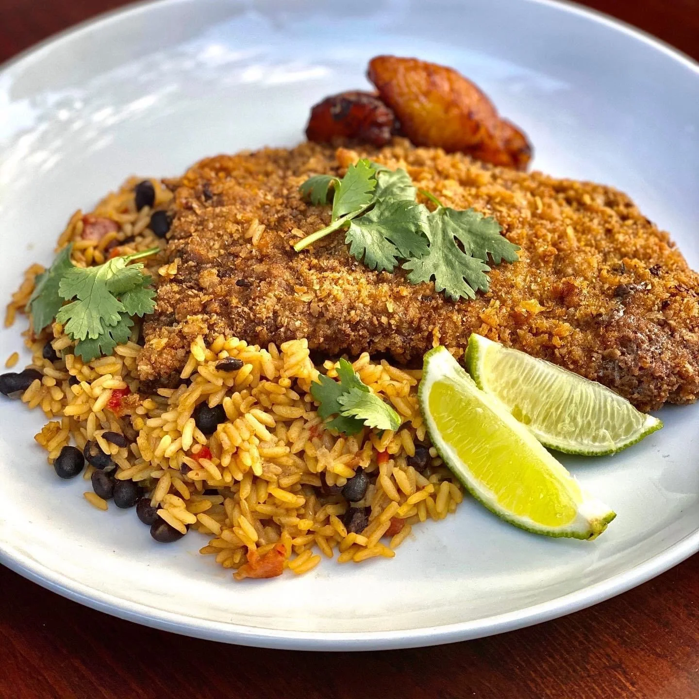 Fried fish fillet with seasoned yellow rice and black beans, garnished with cilantro and lime wedges, served with roasted potato wedges on a white plate.