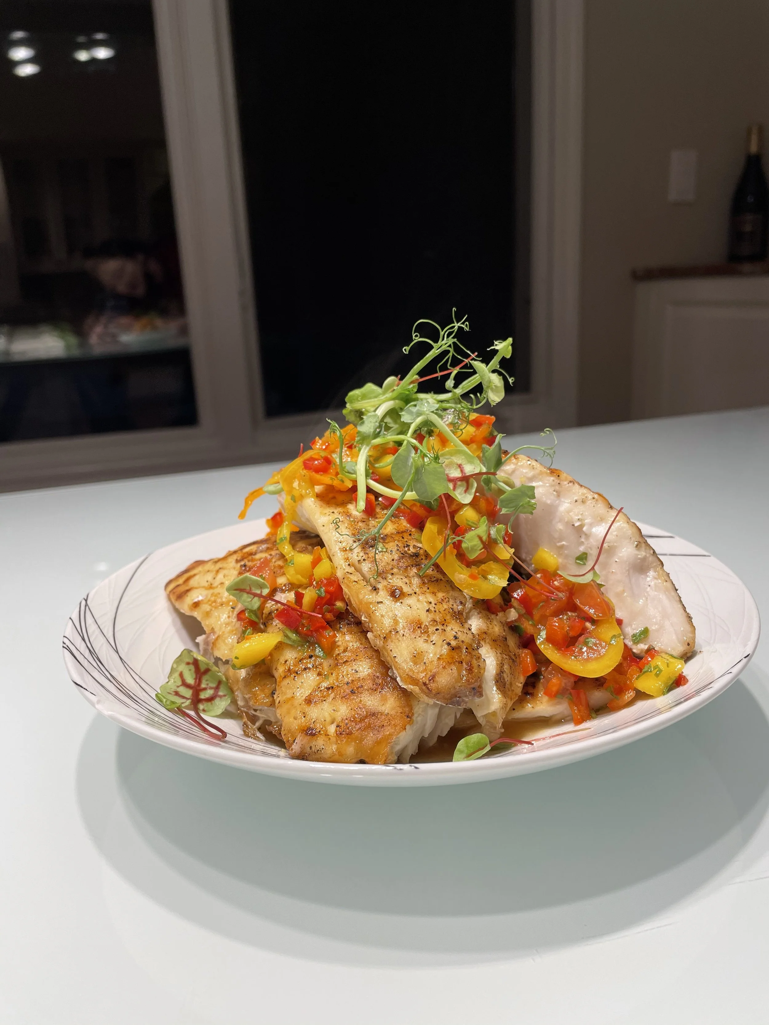 A plated dish of grilled fish topped with a colorful vegetable medley and microgreens, on a white plate.