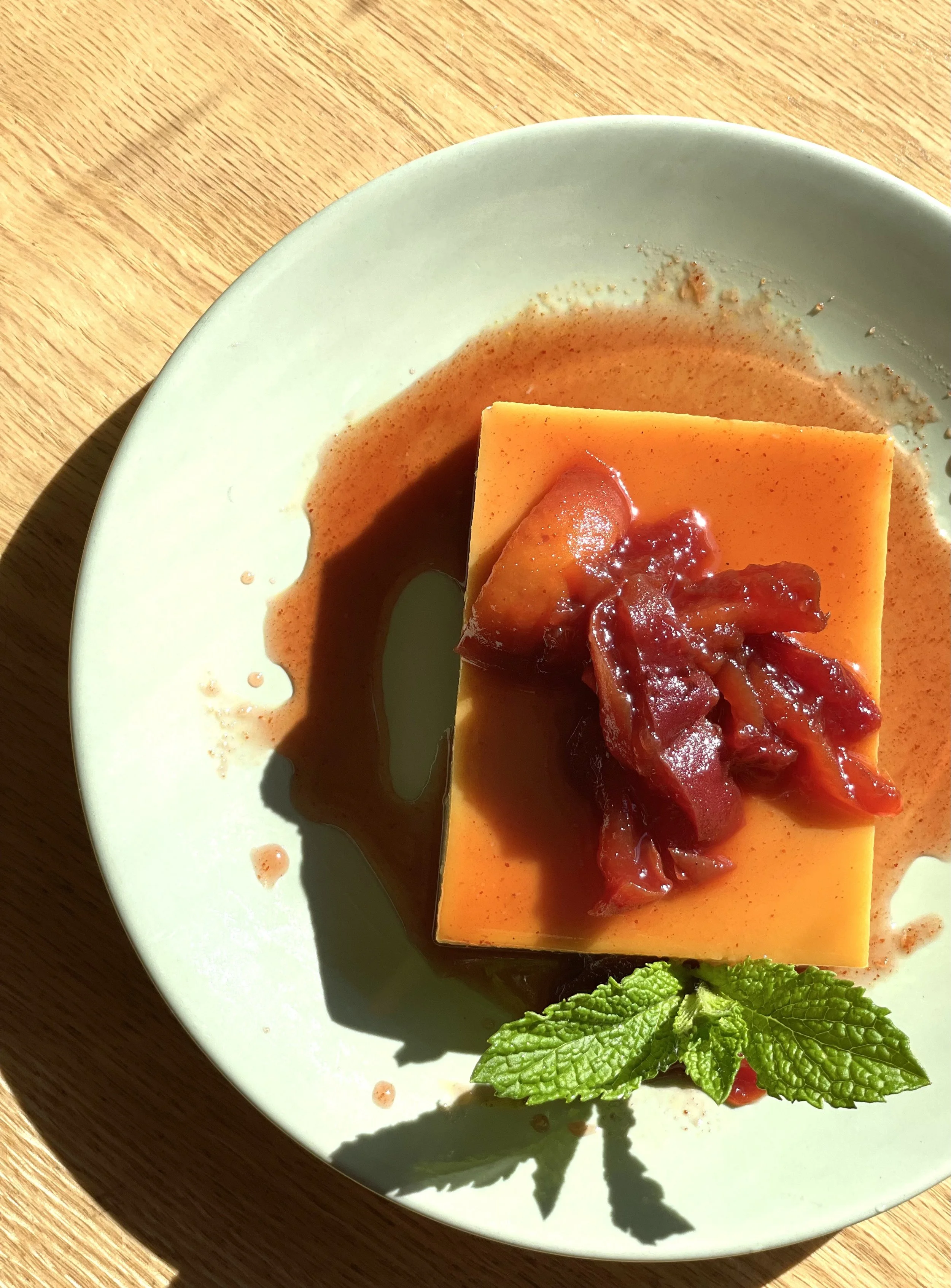 A slice of flan topped with caramelized fruit and sauce, garnished with mint leaves on a green plate.