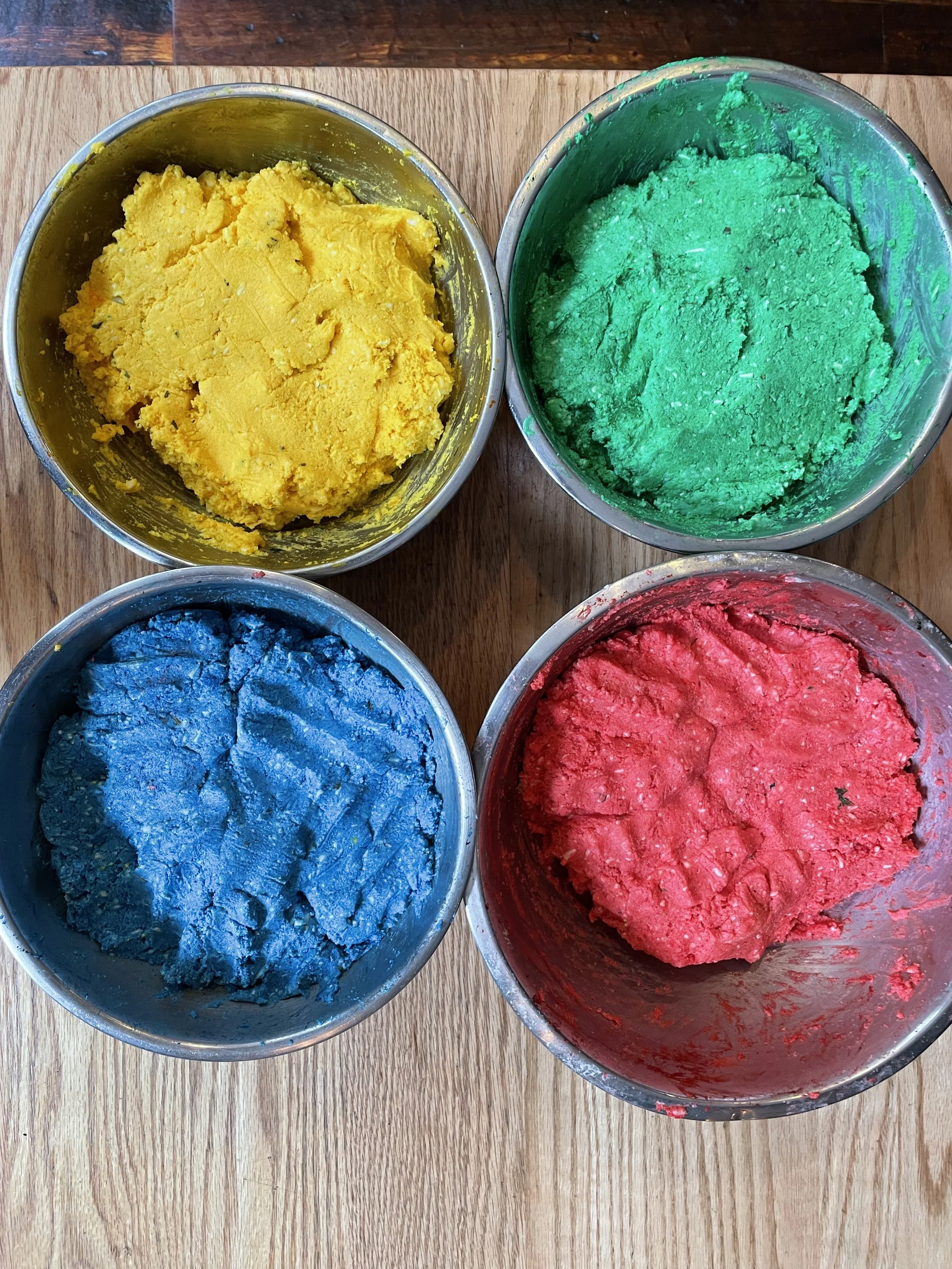 Four metal bowls filled with colored doughs - yellow, green, blue, and red - on a wooden surface.