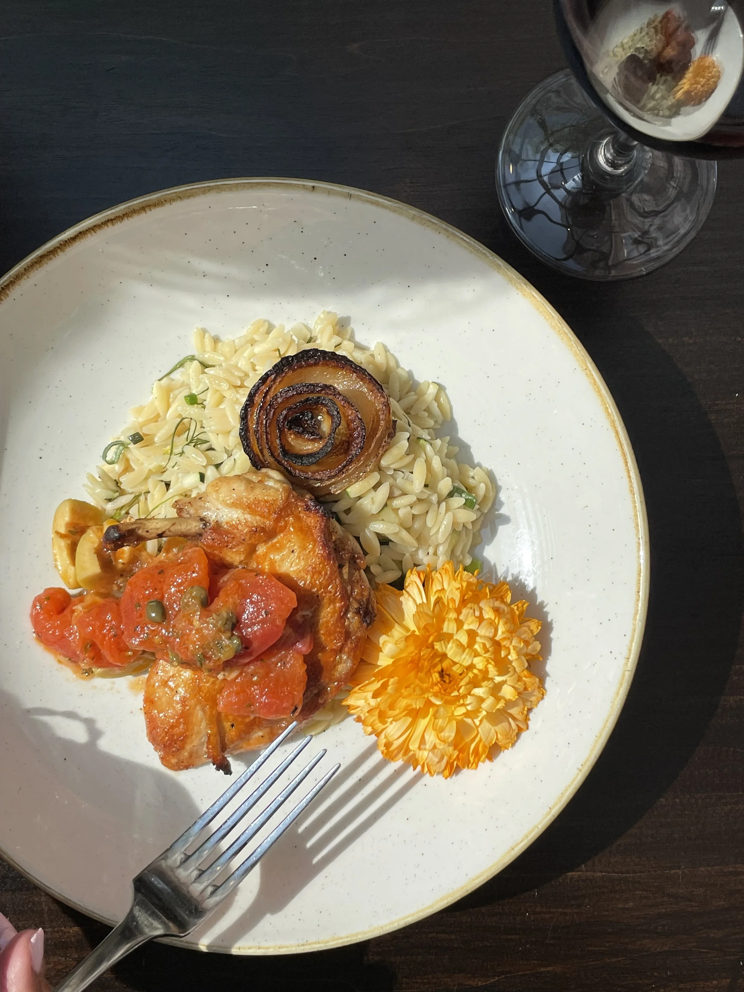 Plate with roasted chicken topped with tomato sauce, rice with green herbs, caramelized onion roll, and orange edible flower, alongside a glass of red wine on a dark wooden table.