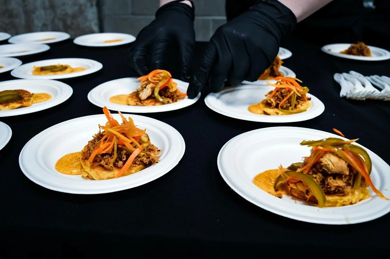 Someone wearing black gloves serving shredded meat tacos topped with shredded carrots and peppers on white plates arranged on a black table.
