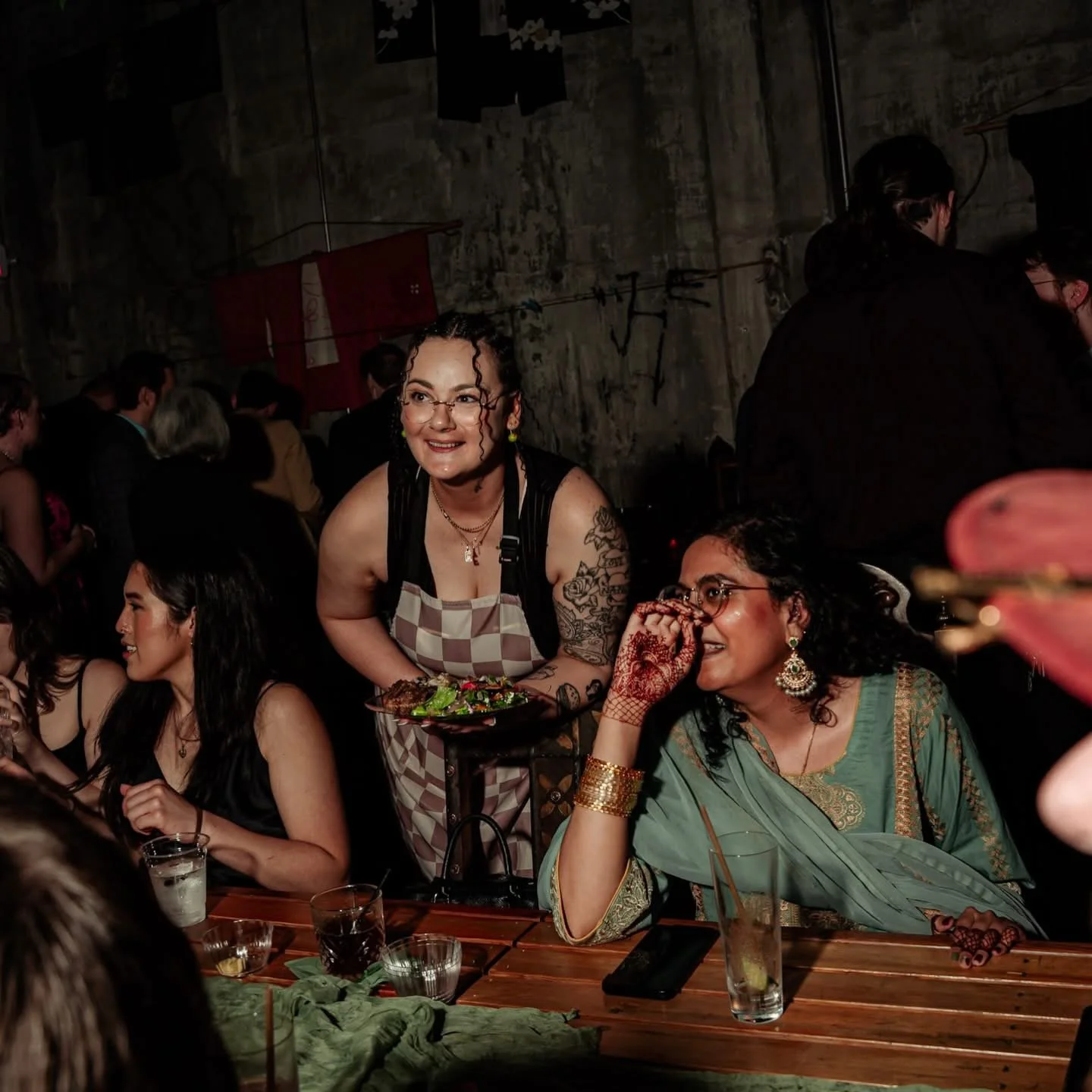 Group of women with Chef Jessica Baston at a social gathering or party, seated at a table with drinks, with a woman in an apron serving salad. The setting appears casual and lively, with a rustic background.