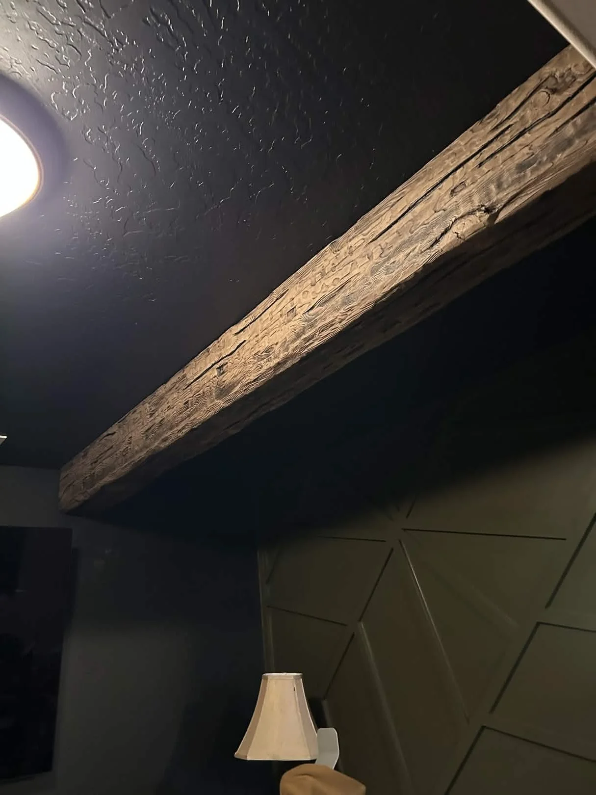 Room corner with black textured wall, wooden beam, ceiling light, section of a lamp, and tiled floor.