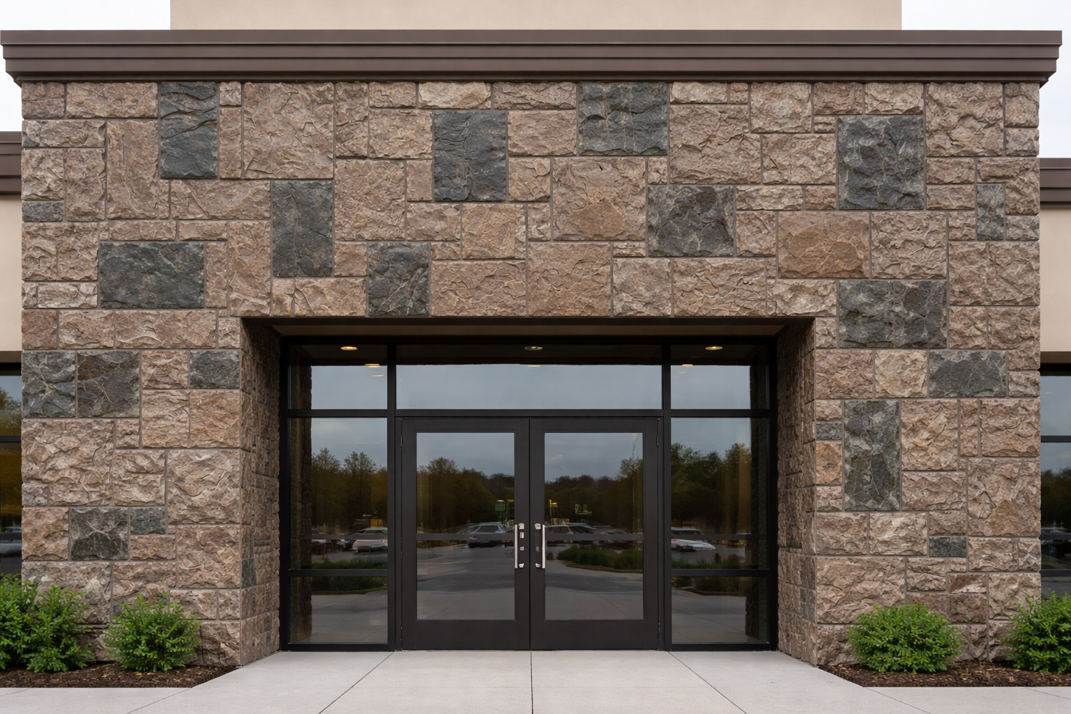 Modern building entrance with stone facade and glass doors, flanked by small green bushes.