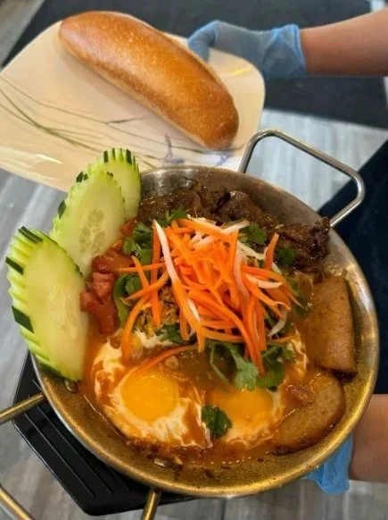 A bowl of spicy meat stew with fried eggs, sliced cucumbers, shredded carrots, and cilantro, accompanied by a piece of bread.