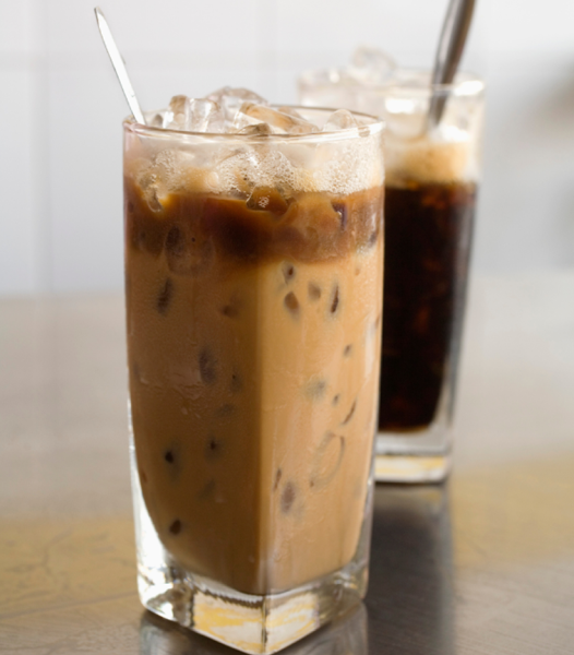 Two glasses of iced coffee, one with milk and the other dark, on a wooden surface with a white tiled background.