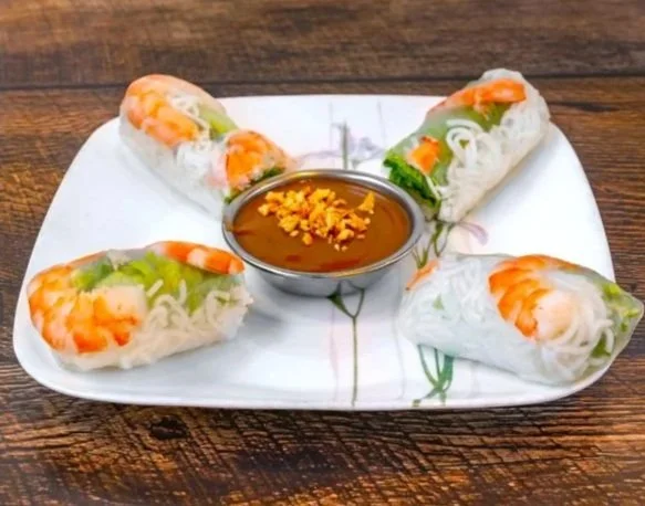 Four Vietnamese spring rolls with shrimp and vegetables on a white plate, served with a bowl of dipping sauce garnished with crushed peanuts, on a wooden table.
