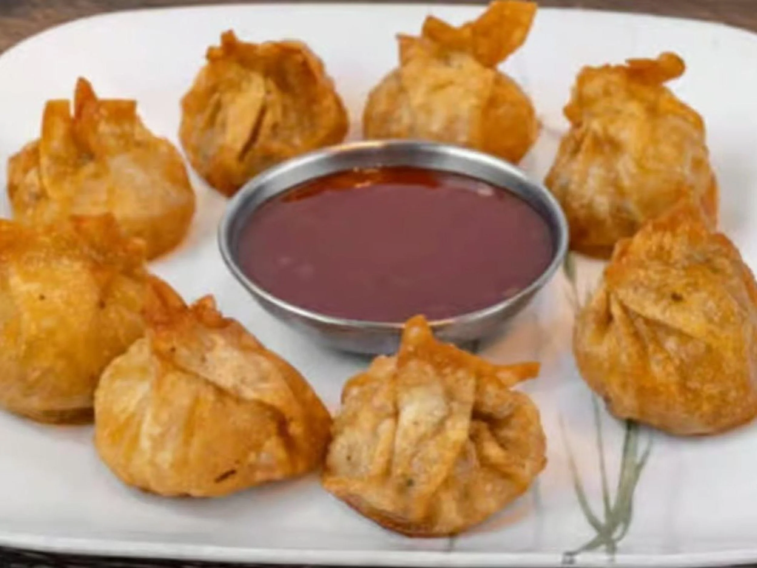 Eight crispy fried samosas arranged on a white plate with a metal bowl of dark red dipping sauce in the center.