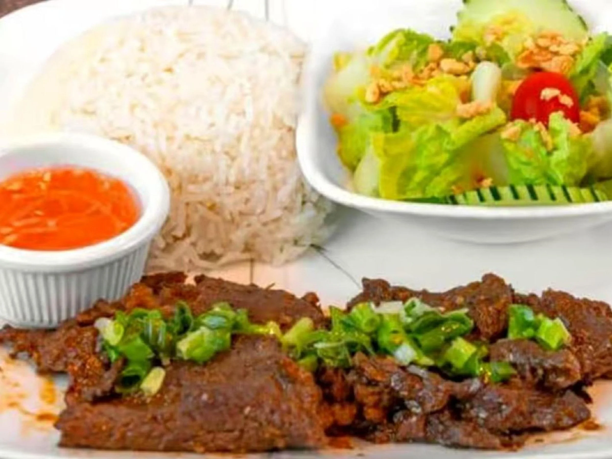 Plate with grilled meat topped with chopped green onions, a bowl of orange-colored dipping sauce, a serving of rice, and a small bowl of salad with lettuce, cherry tomato, and crushed peanuts.