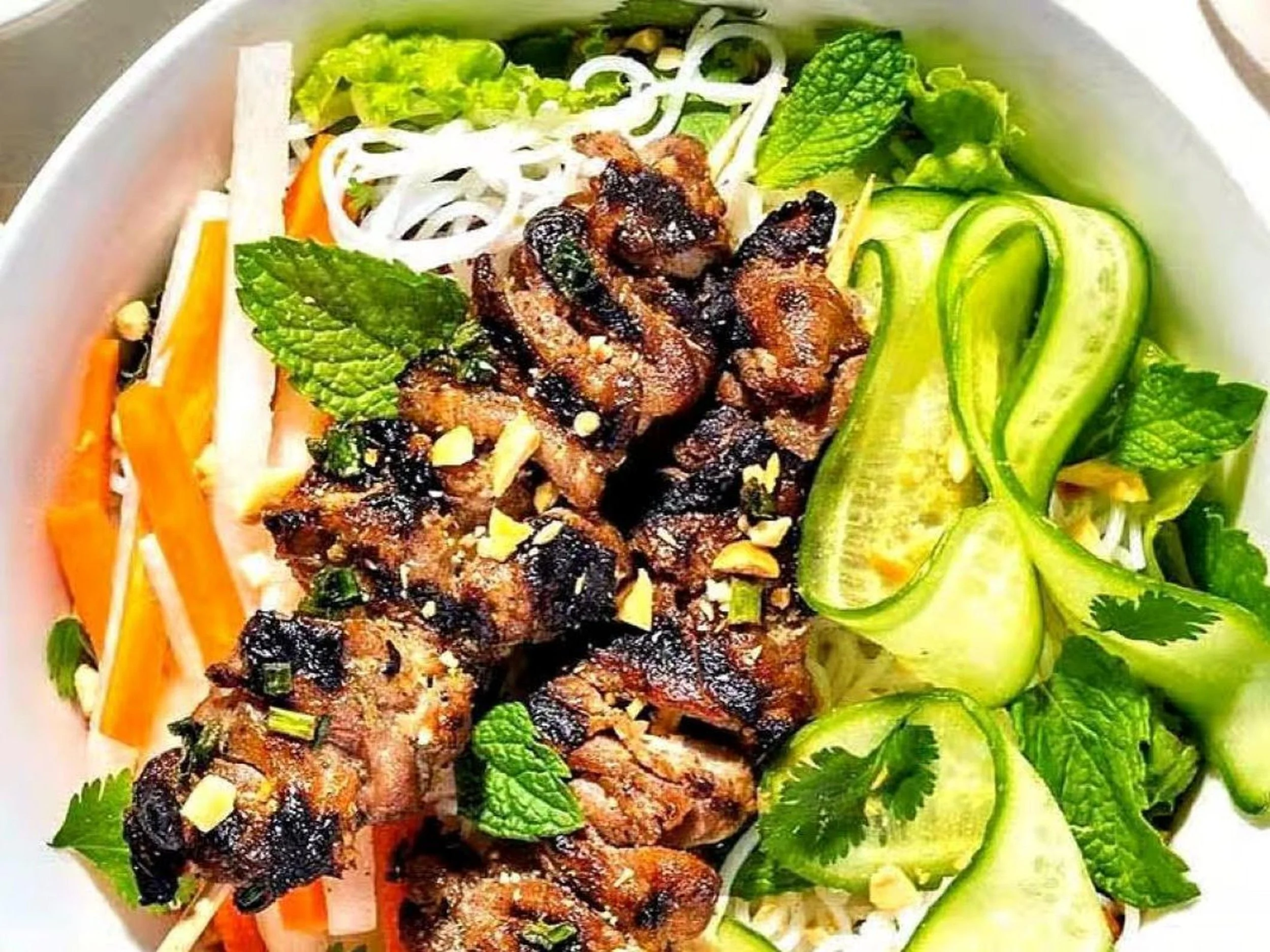 Skewered grilled meat with fresh vegetables and herbs, including sliced cucumber, carrots, lettuce, mint leaves, and bean sprouts.