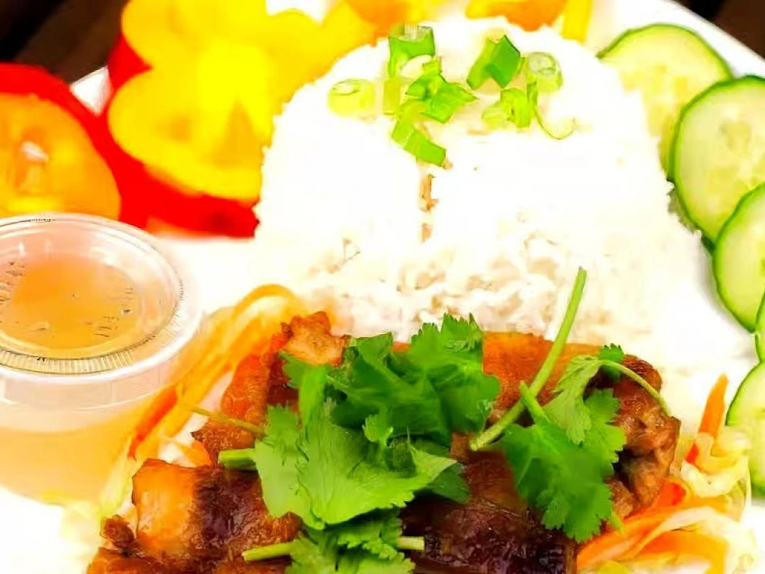 Plate of Thai food with cilantro, white rice, sliced cucumbers, sliced tomatoes, a small container of yellow sauce, grilled meat, shredded carrots, and onions.