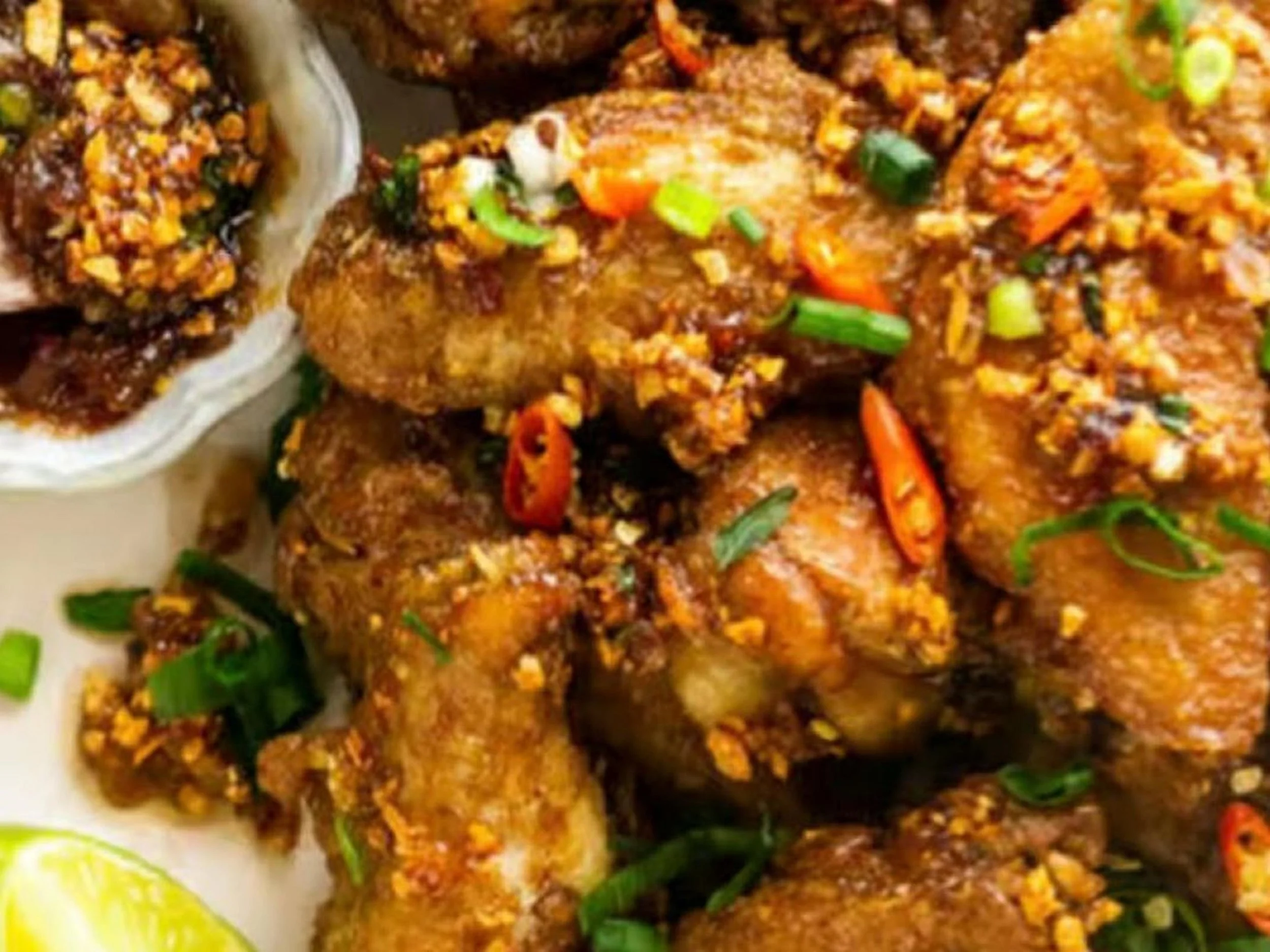 Close-up of fried chicken pieces garnished with chopped green onions, red chili slices, and crushed peanuts, served with a lemon wedge on the side.