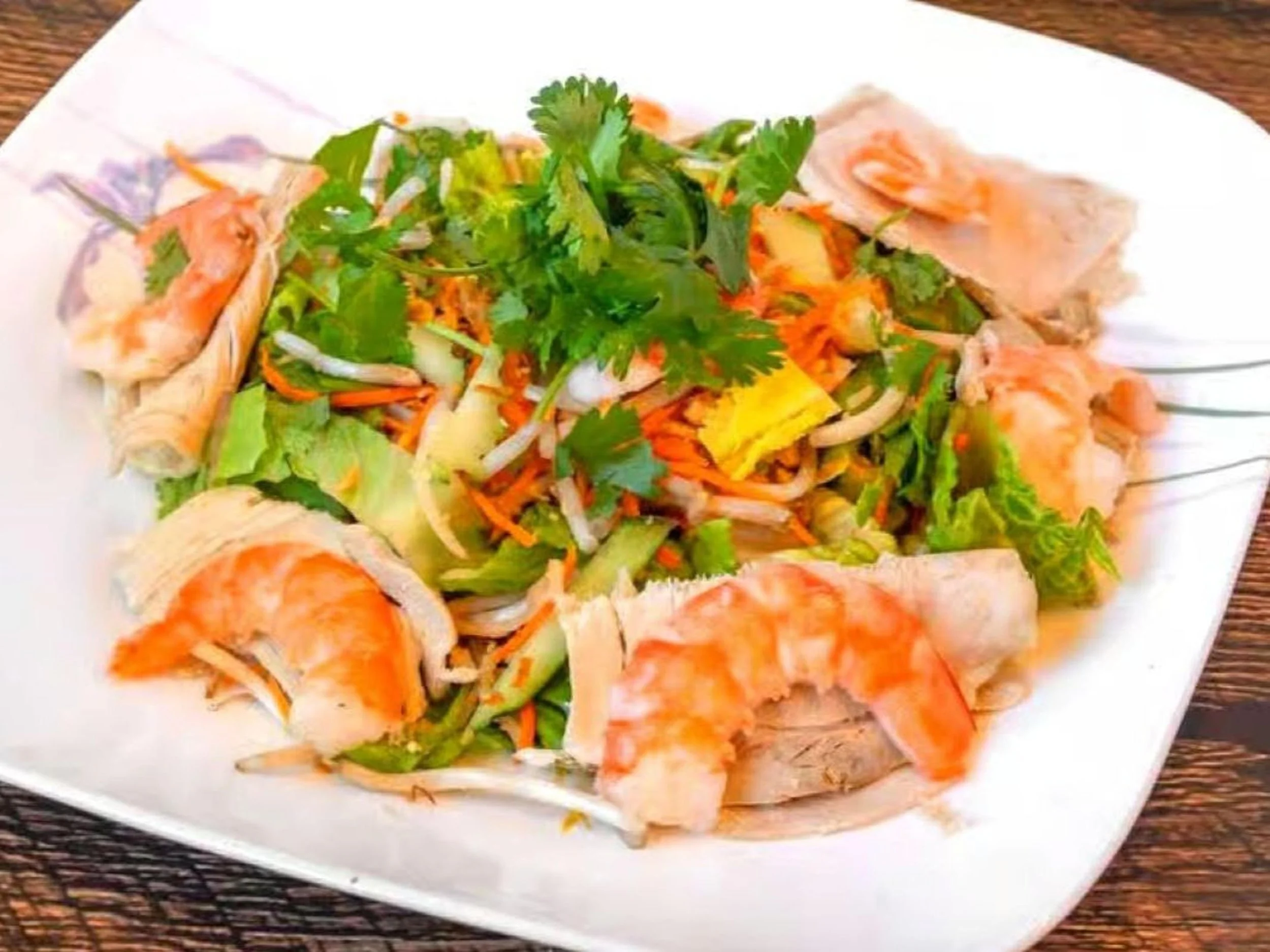 Salad with shrimp, lettuce, shredded carrots, cilantro, and sliced chicken on a white plate on a wooden table.