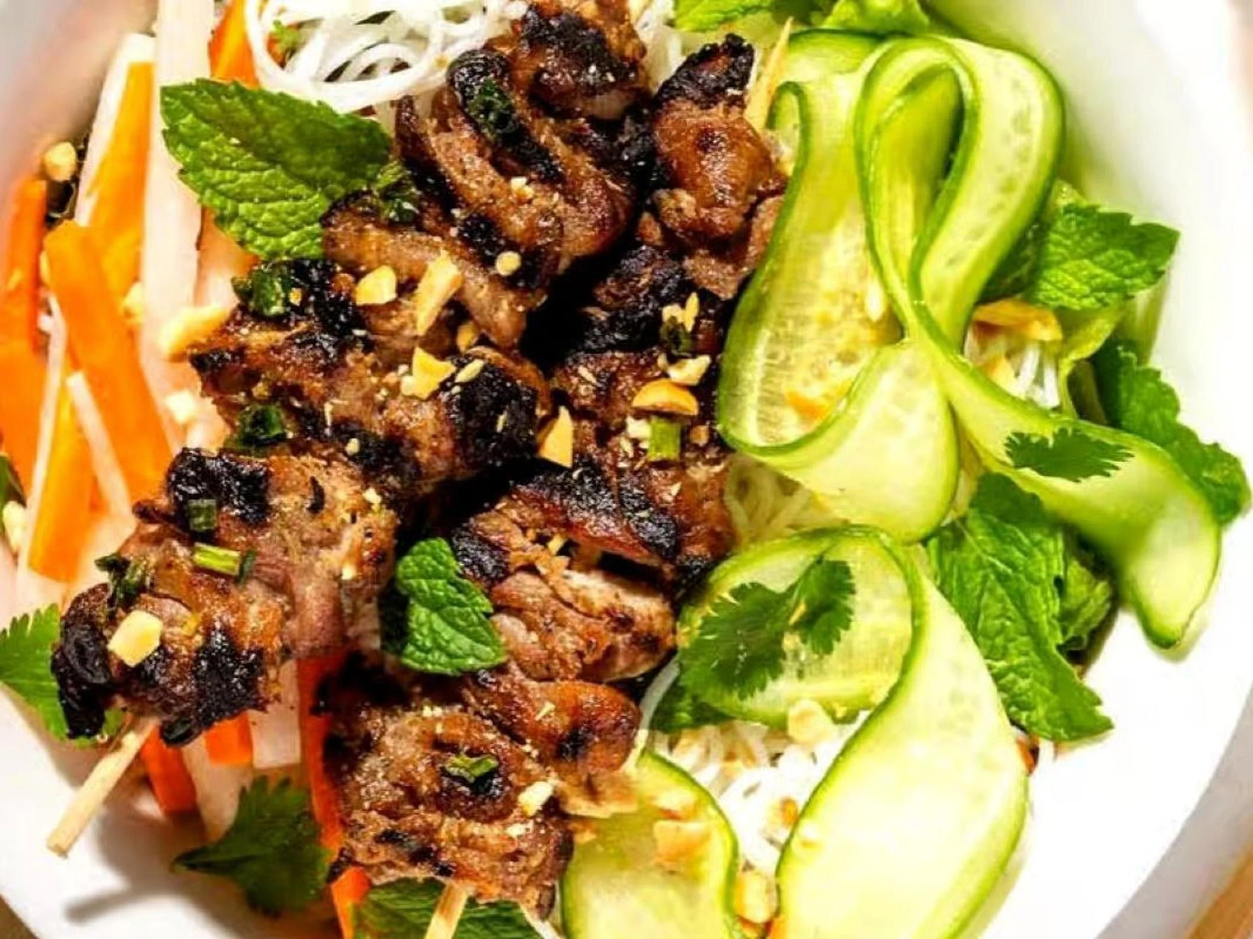 Fresh grilled meat skewers with chopped garlic and herbs, served on a bed of sliced vegetables including cucumber ribbons, carrots, and bean sprouts, garnished with mint and cilantro.