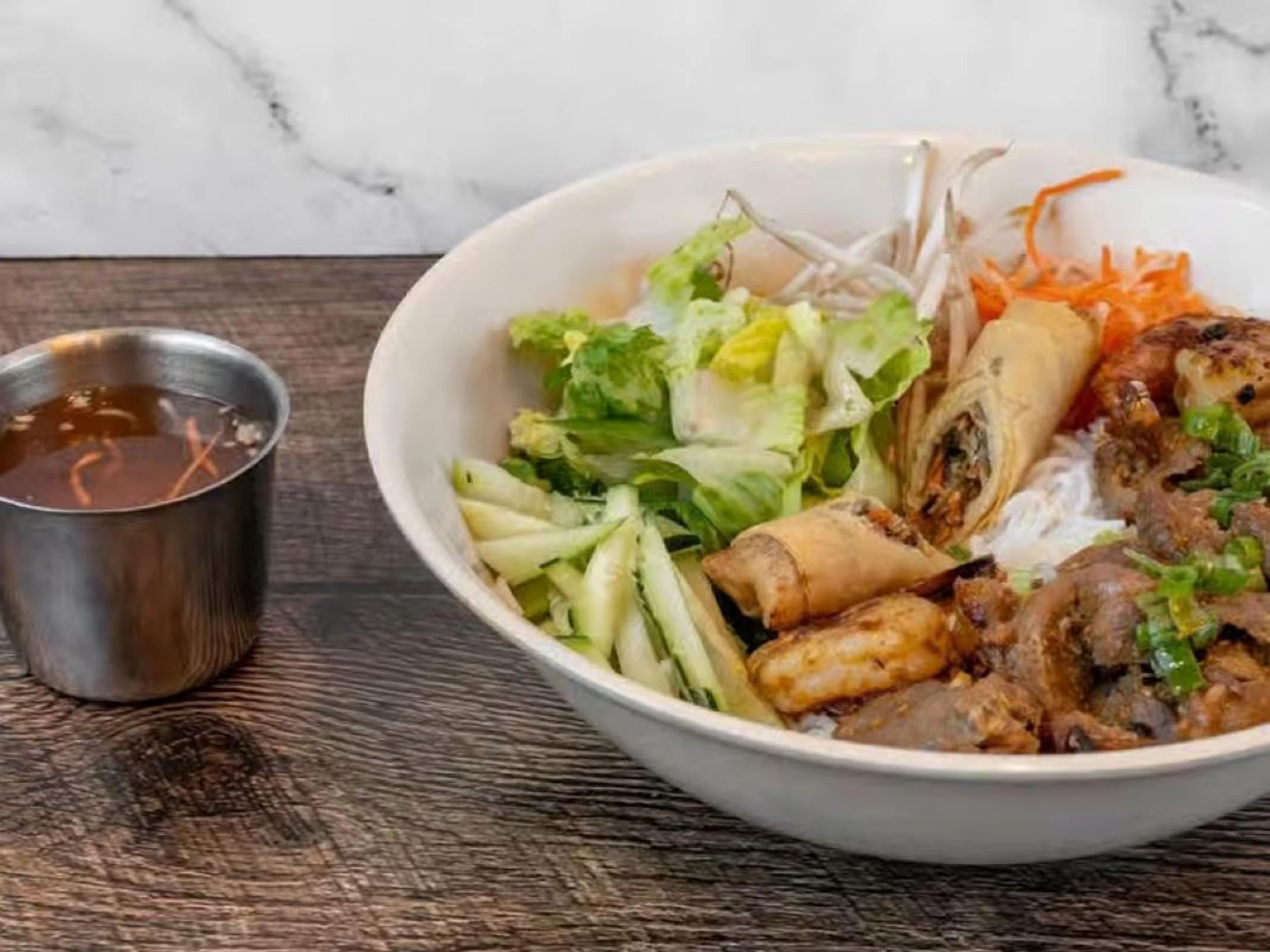 A bowl of Asian cuisine with spring rolls, chopped vegetables, and grilled chicken on a wooden table, with a small cup of dipping sauce on the side.
