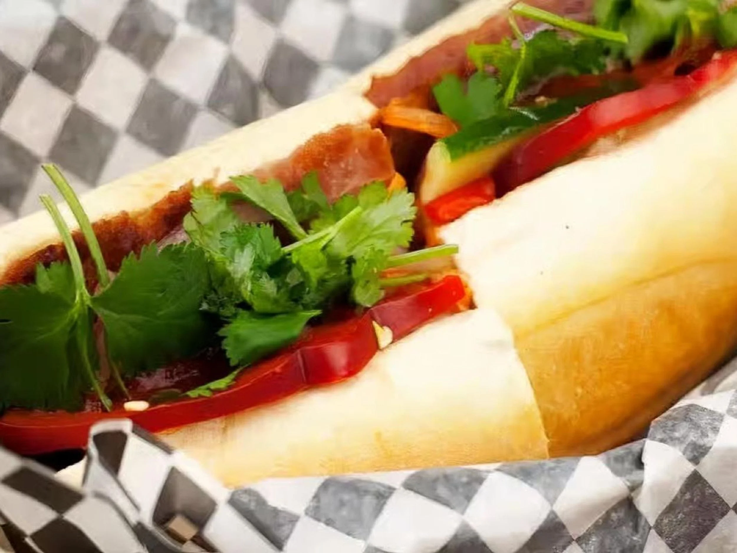 Close-up of a sandwich with sliced bread, fresh cilantro, and sliced red bell peppers inside on a checkered paper.