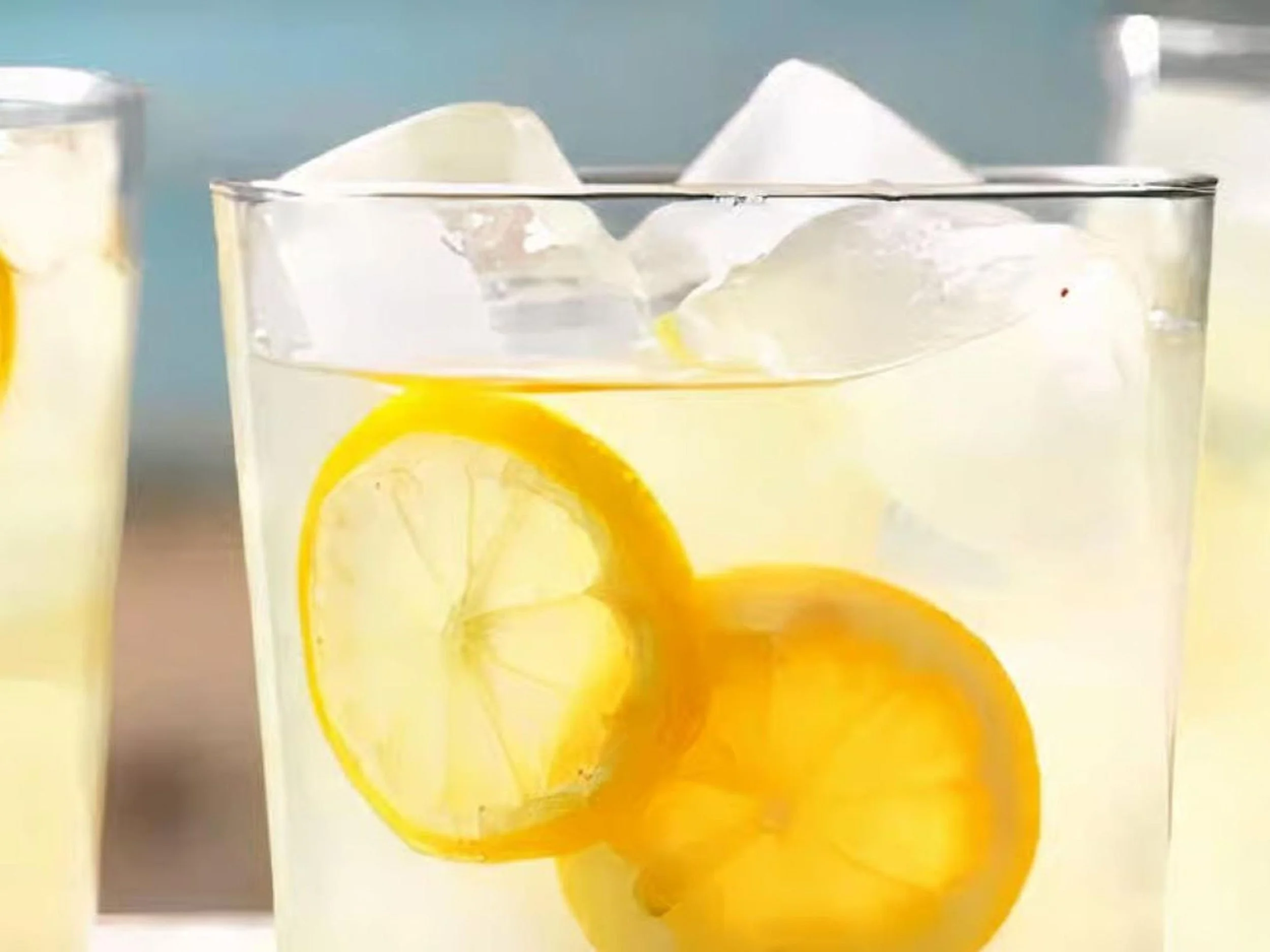 A glass of lemonade with lemon slices and ice cubes, with more glasses of lemonade in the background.