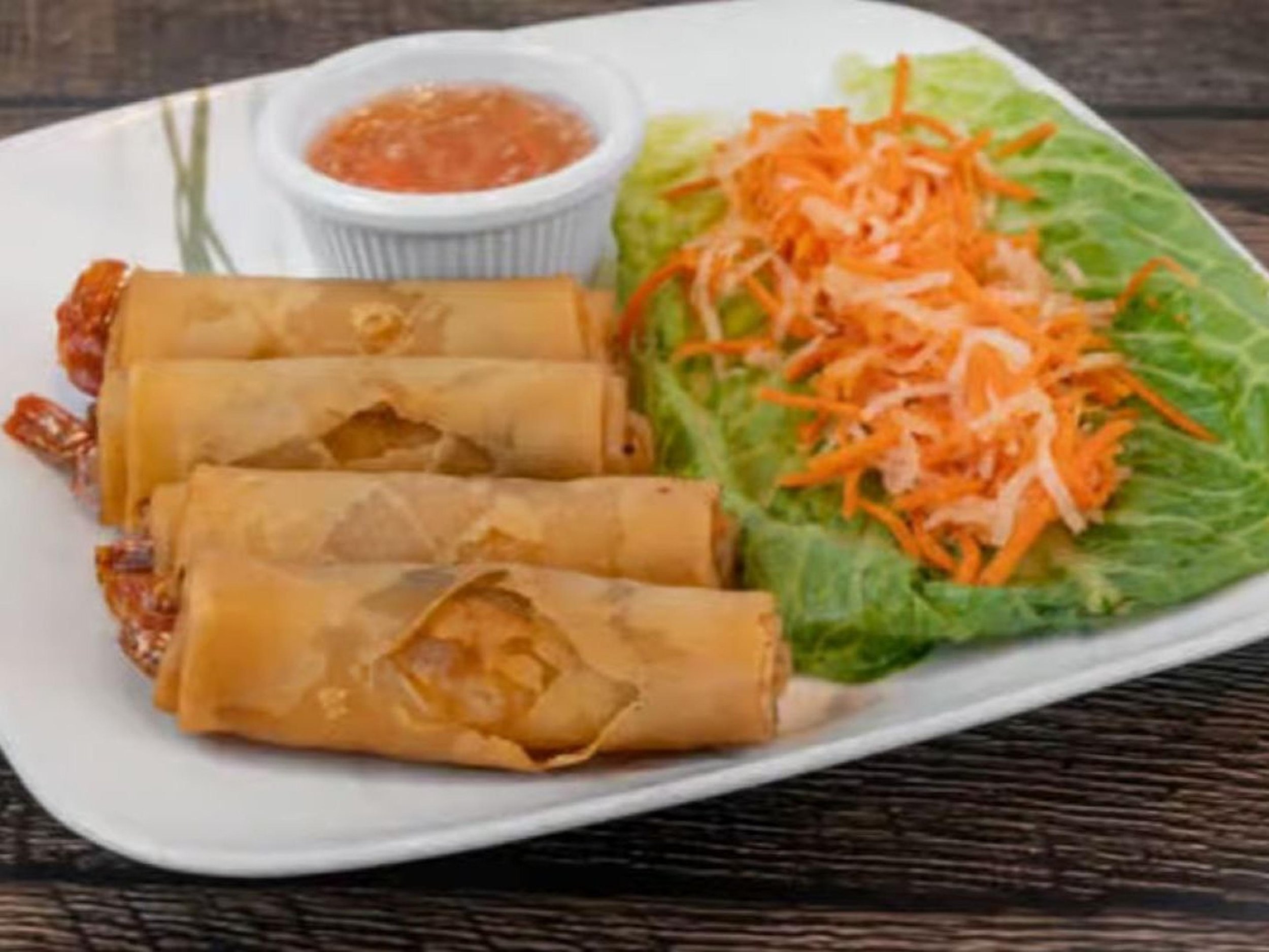 Plate of three spring rolls with shredded carrots and lettuce, accompanied by a small bowl of dipping sauce on a white plate.
