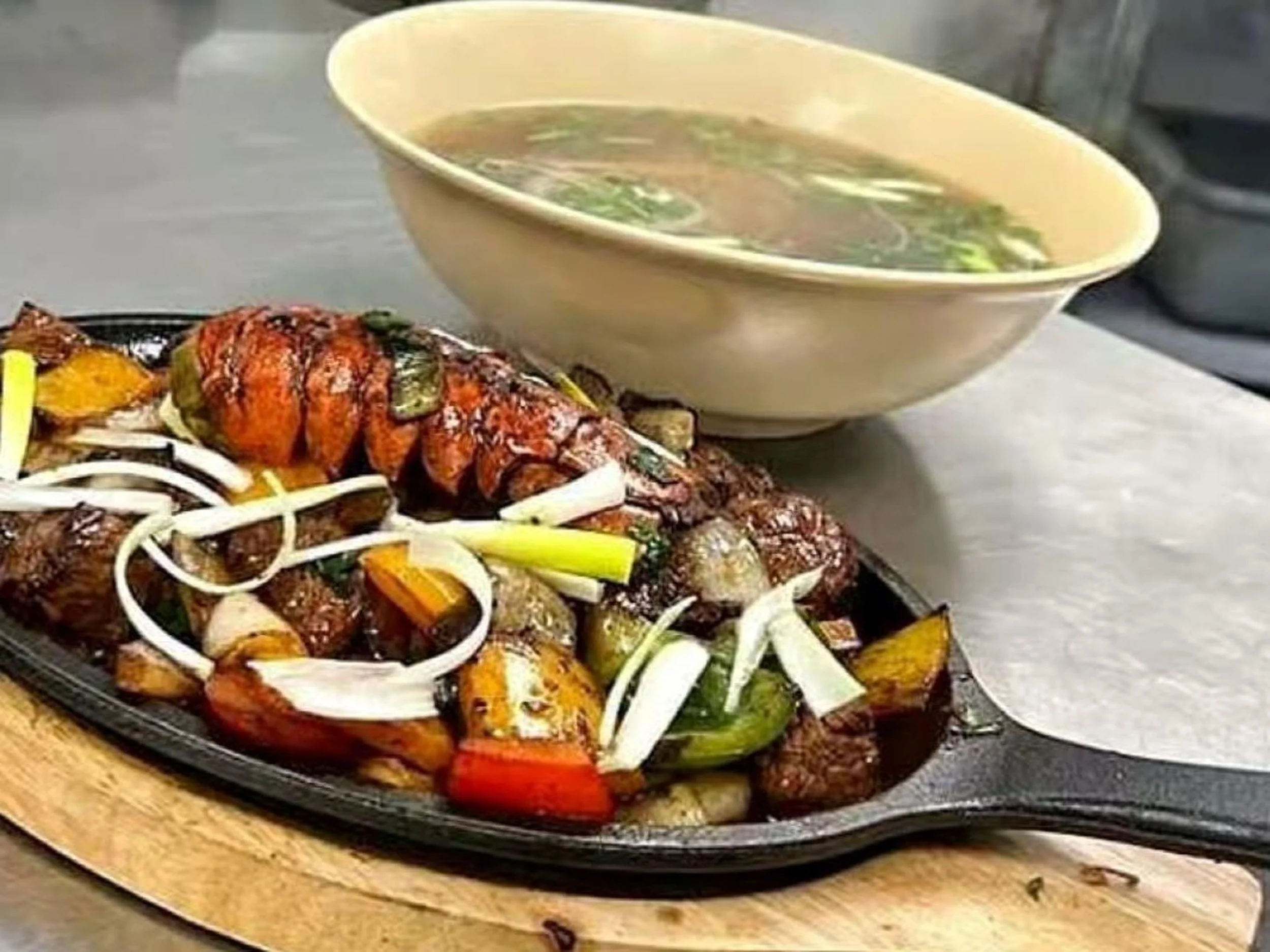 Grilled lobster tail with vegetables served on a cast iron skillet, accompanied by a bowl of soup with green onions.