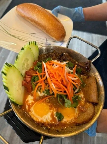 A plate of spicy beef noodle stew with vegetables, fried egg, cucumber slices, and spring roll, served with a bread roll.