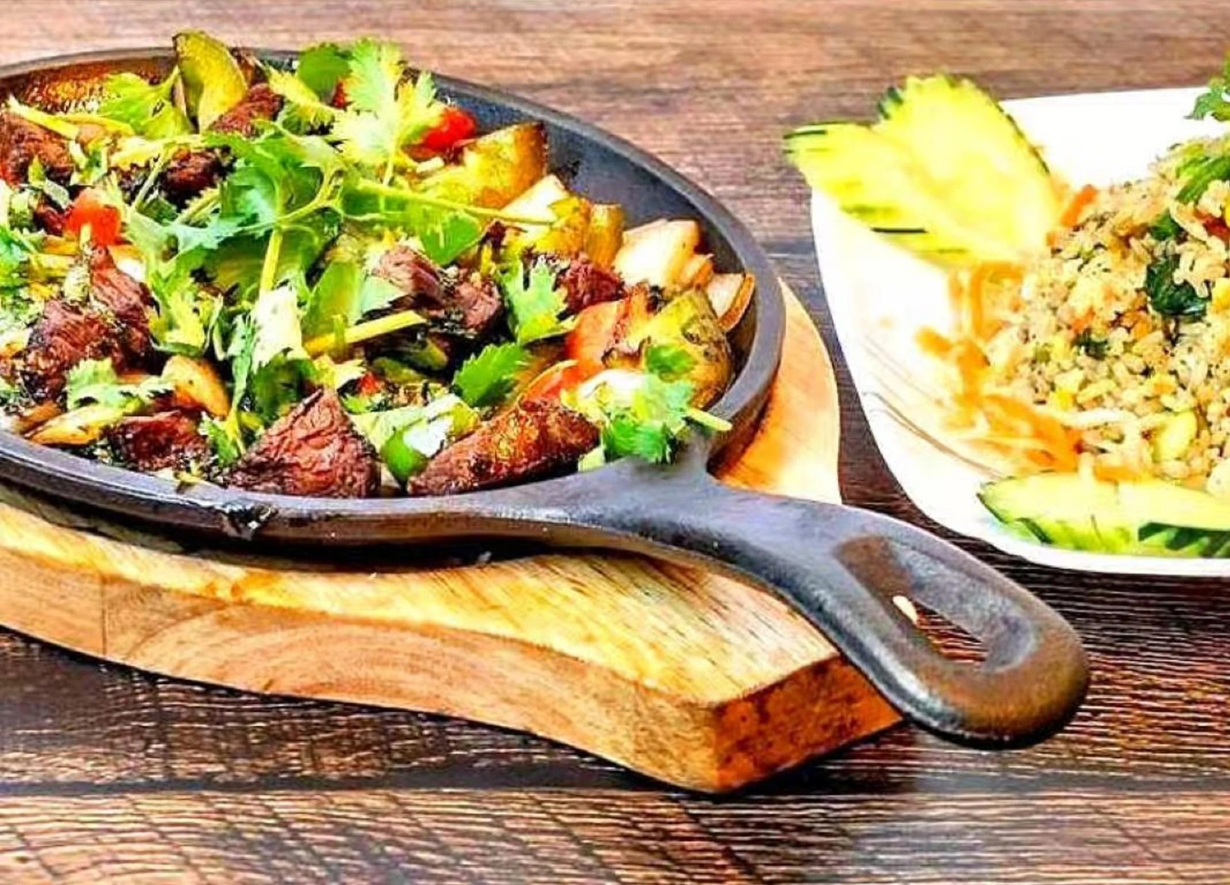 A serving of beef stir-fry with vegetables, served on a black skillet on a wooden board, with a side of rice and cucumber slices.