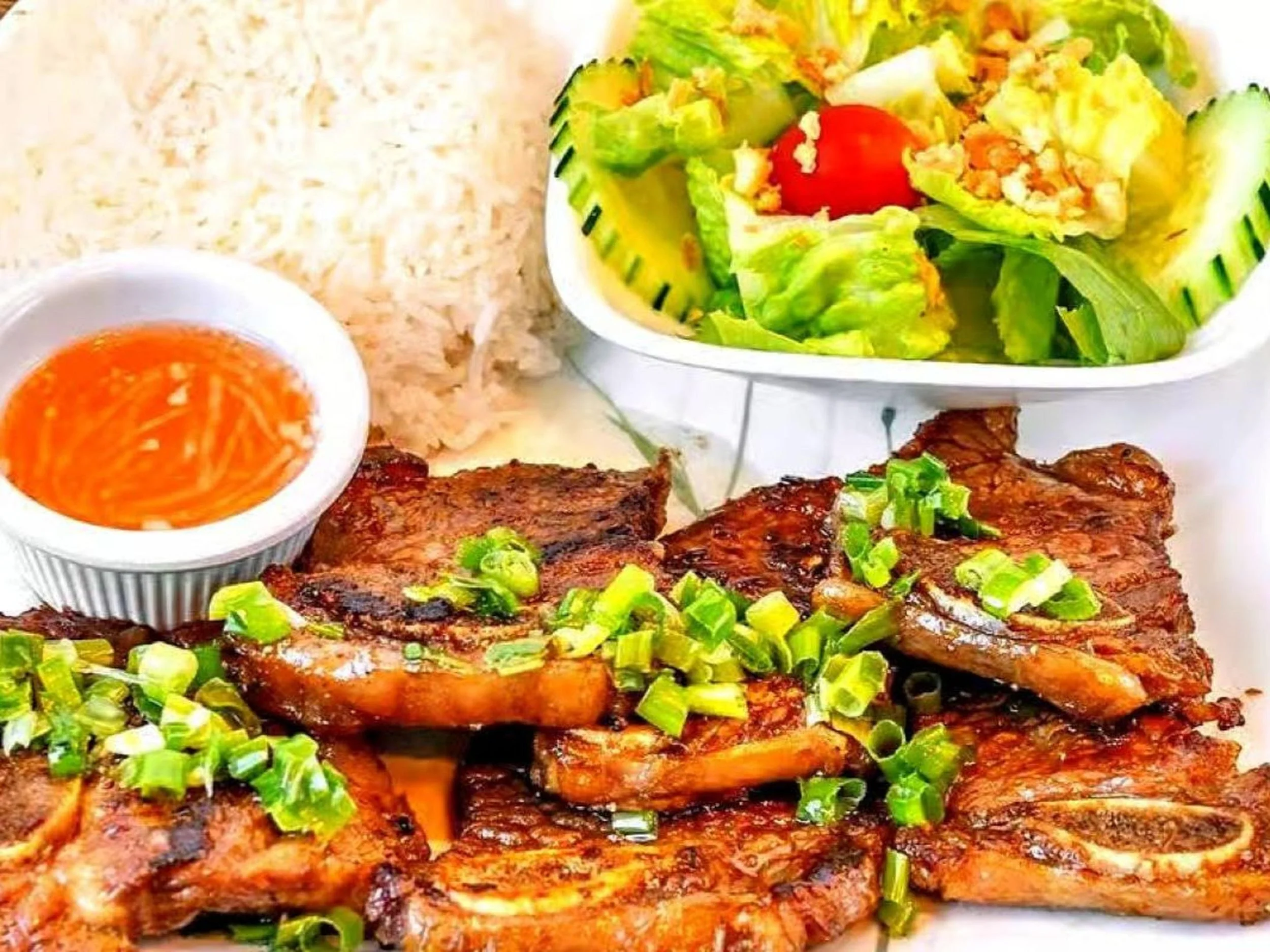 Grilled pork chops garnished with chopped green onions served with rice, a small bowl of dipping sauce, a side of salad with lettuce, cherry tomato, and cucumber slices in a white bowl.